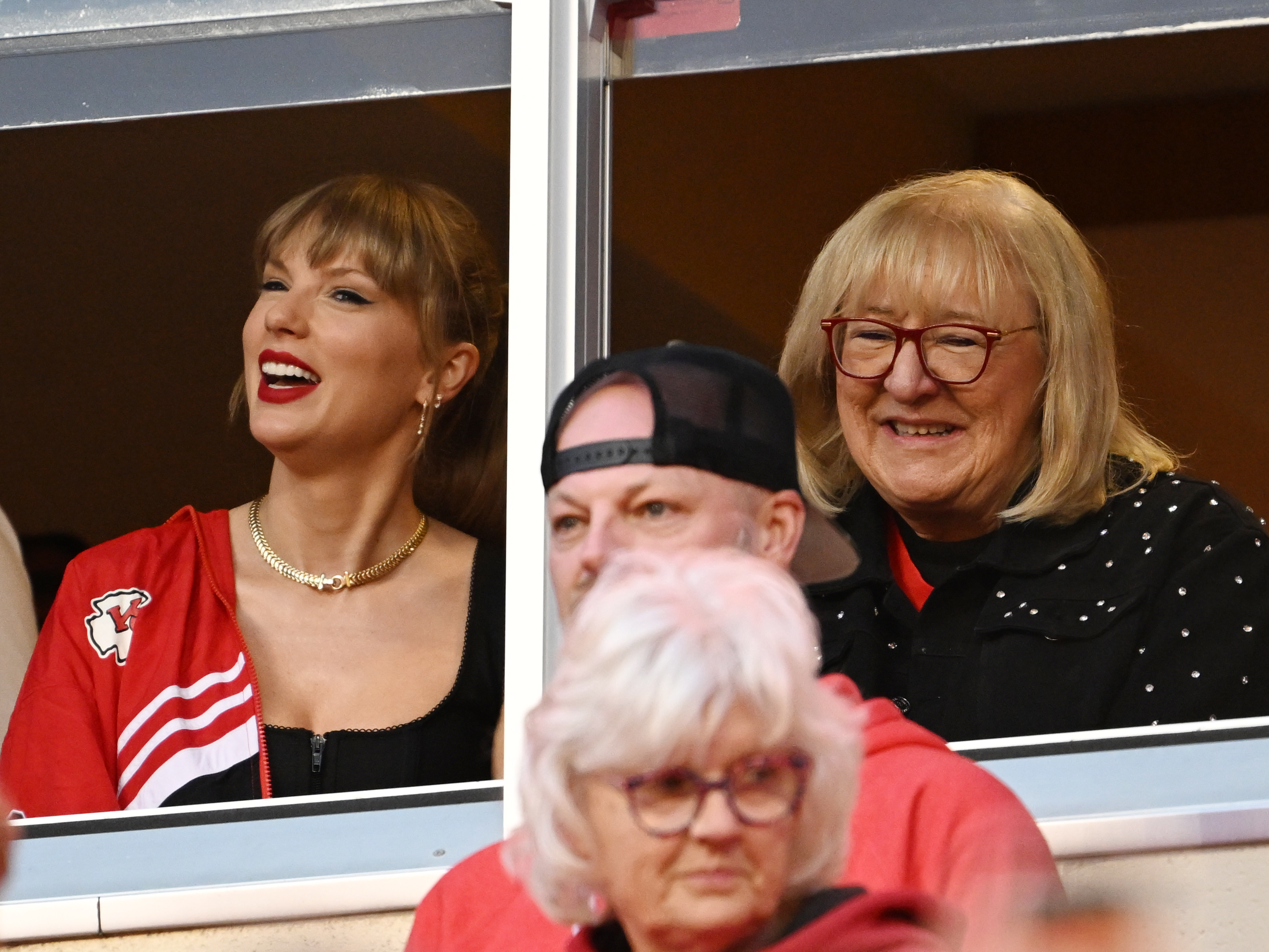 Taylor Swift, wearing a red Kansas City Chiefs jacket over a black top, stands beside Donna Kelce, in a black studded jacket, as they smile and watch the game from a private suite at GEHA Field at Arrowhead Stadium, on October 12, 2023, in Missouri.