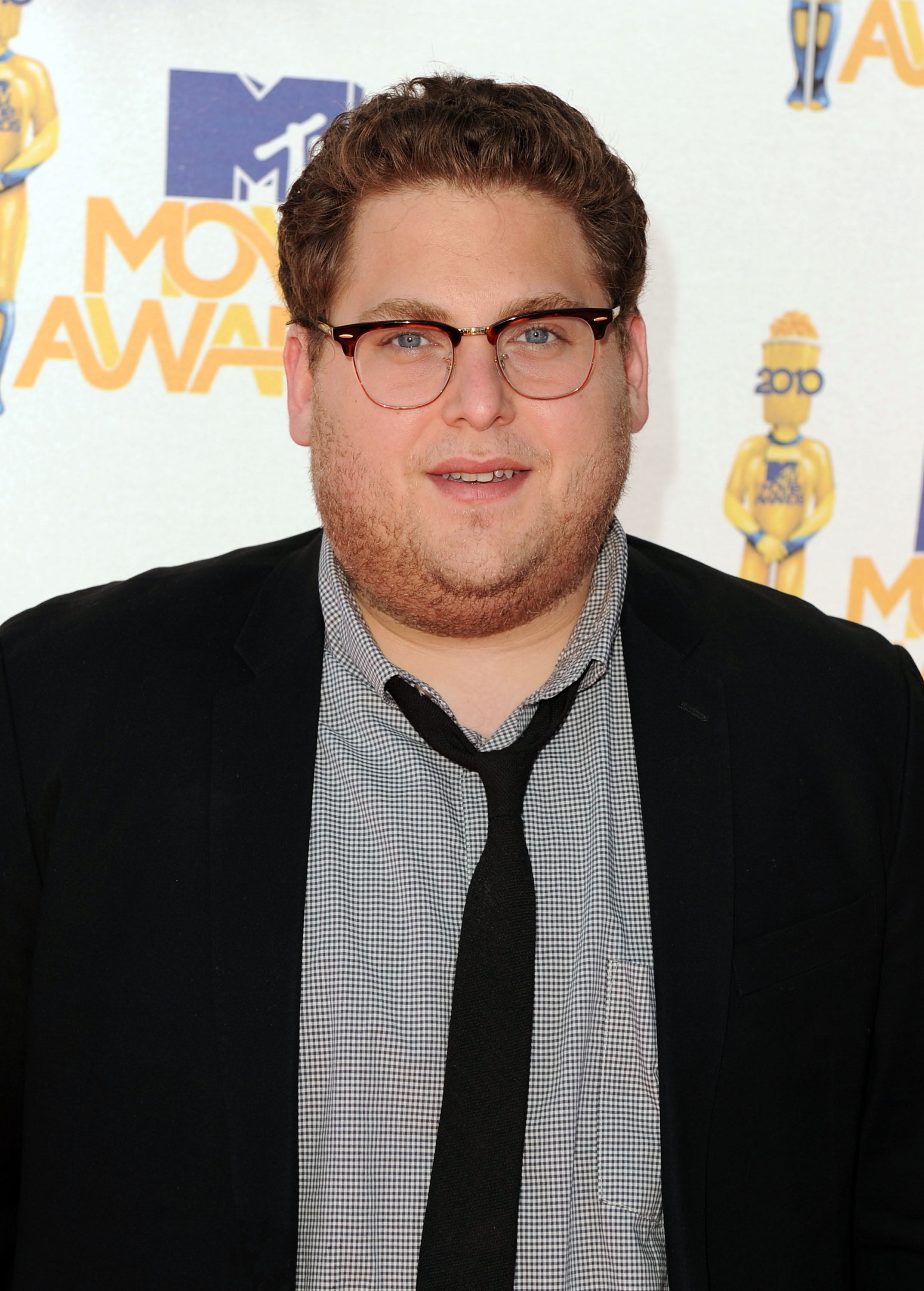 Jonah Hill appears on the red carpet at the 2010 MTV Movie Awards wearing a black blazer over a small checkered shirt, paired with a loosely knotted black tie. He sports short, neatly styled hair and round tortoiseshell glasses for the occasion.