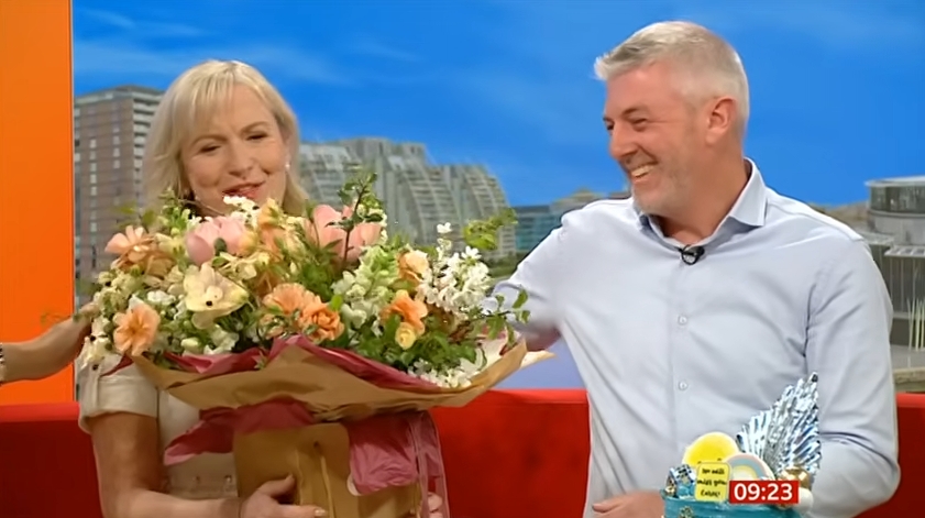 Kirkwood buries her face in the bouquet as Randall looks on with a grin during her emotional last appearance on BBC Breakfast. | Source: YouTube/Weegie Bored