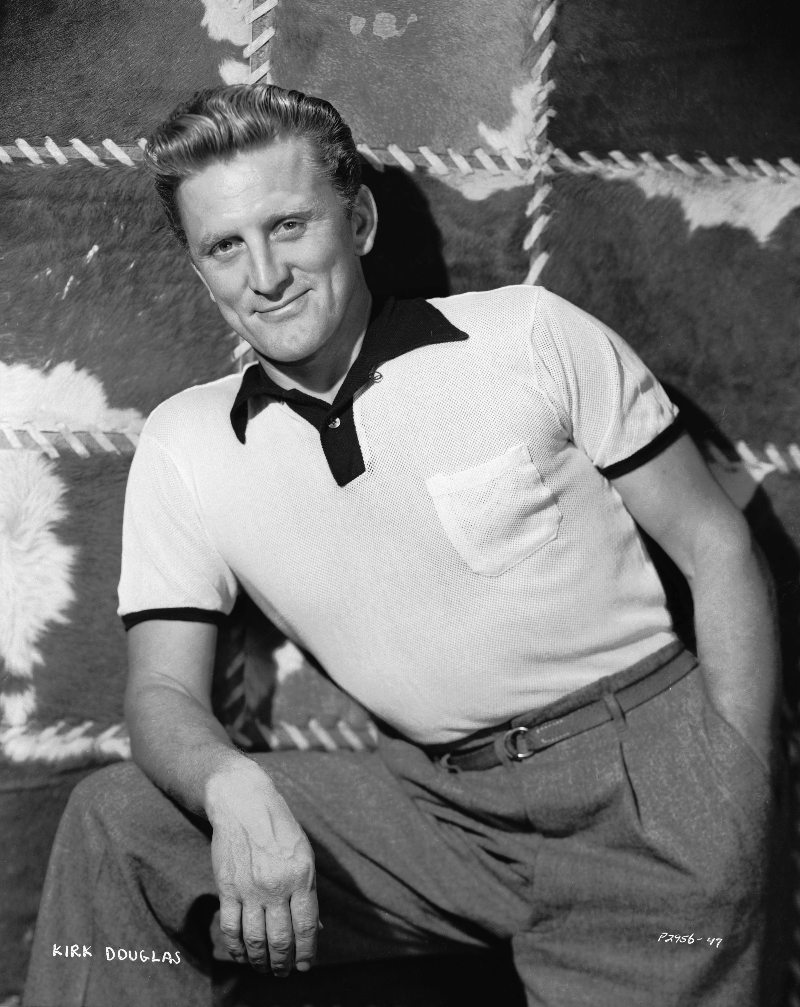  Kirk Douglas posing with one hand in his pocket while resting his other arm on his lap, wearing a black and white t-shirt and gray pants in 1950. / Source: Getty Images