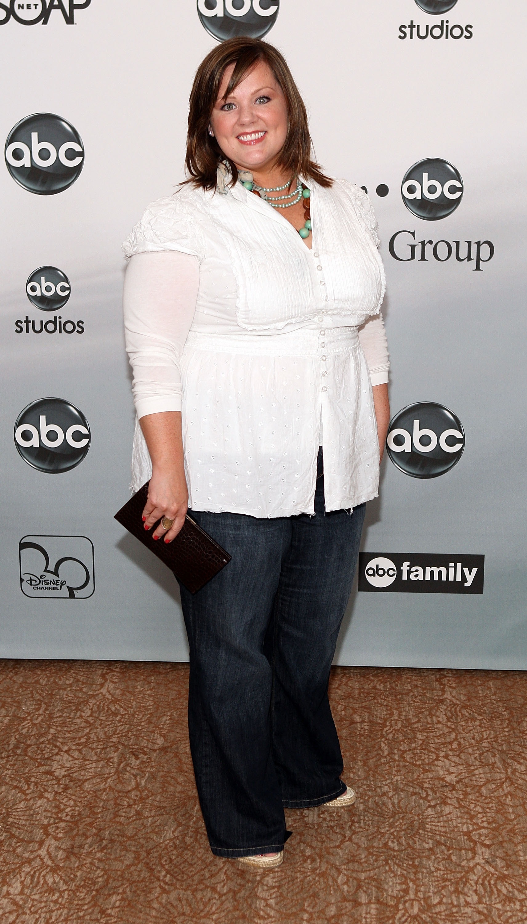 Appearing cheerful and polished, Melissa McCarthy wears a crisp white blouse paired with dark jeans and layered turquoise beads. With a clutch in hand and a bright smile, she brings an easy, welcoming charm to the event backdrop.