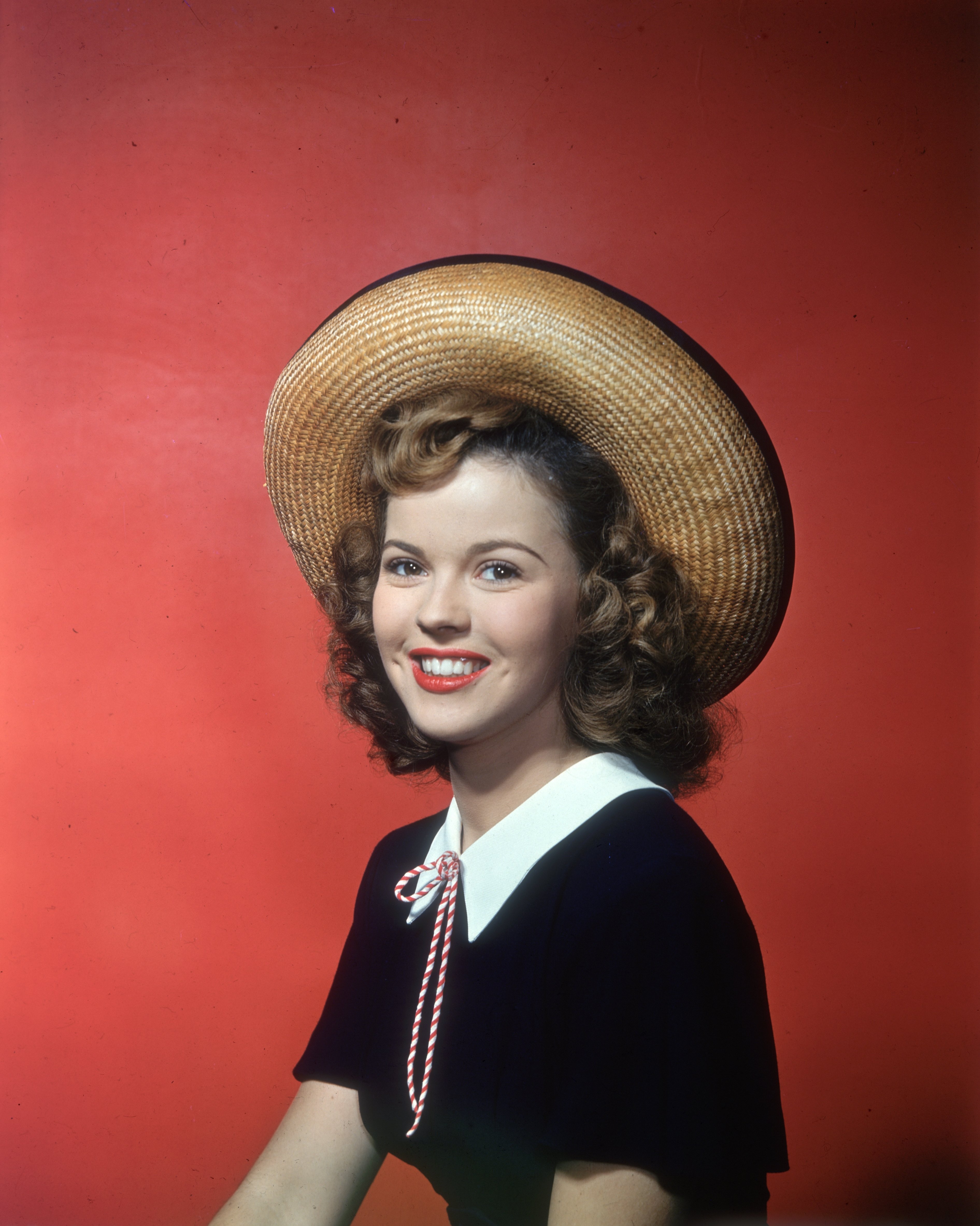  Smiling studio portrait of American actor Shirley Temple, sitting in front of a red backdrop, wearing a straw hat and black top with white collar. circa 1945 |Source: Getty Images
