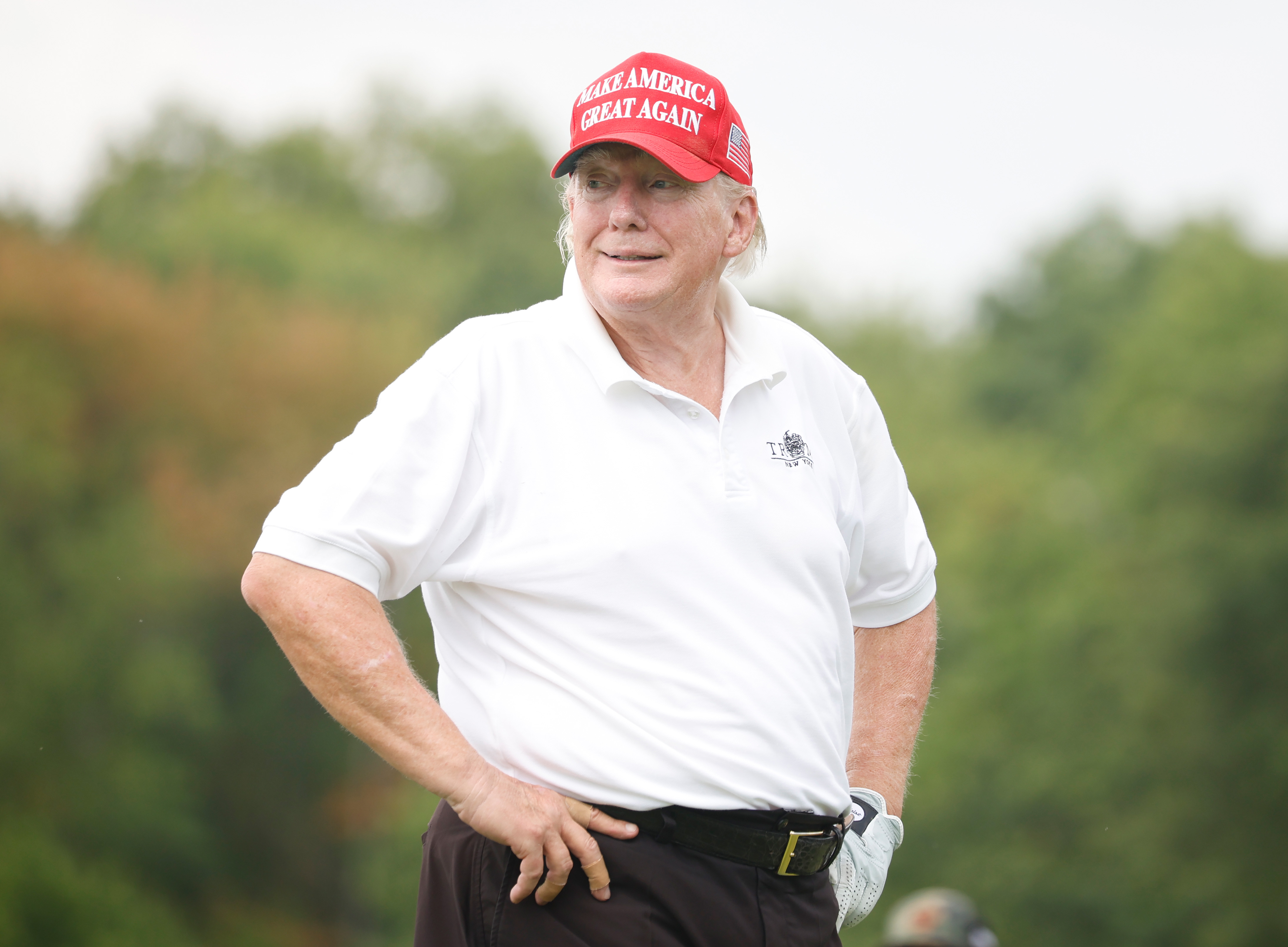 Wearing his signature red cap and white polo, Donald Trump stands on the golf course with hands on his hips. His cheeks and jawline appear noticeably red, contrasting with the pale skin on his neck.