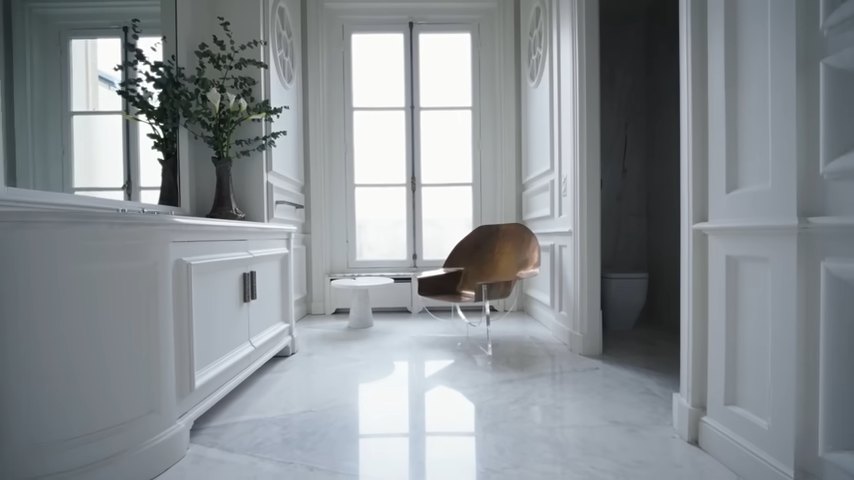 The guest bathroom features a polished white marble floor, a classic white vanity, and a striking, sculptural copper chair by the window. | Source: YouTube/@Archdigest