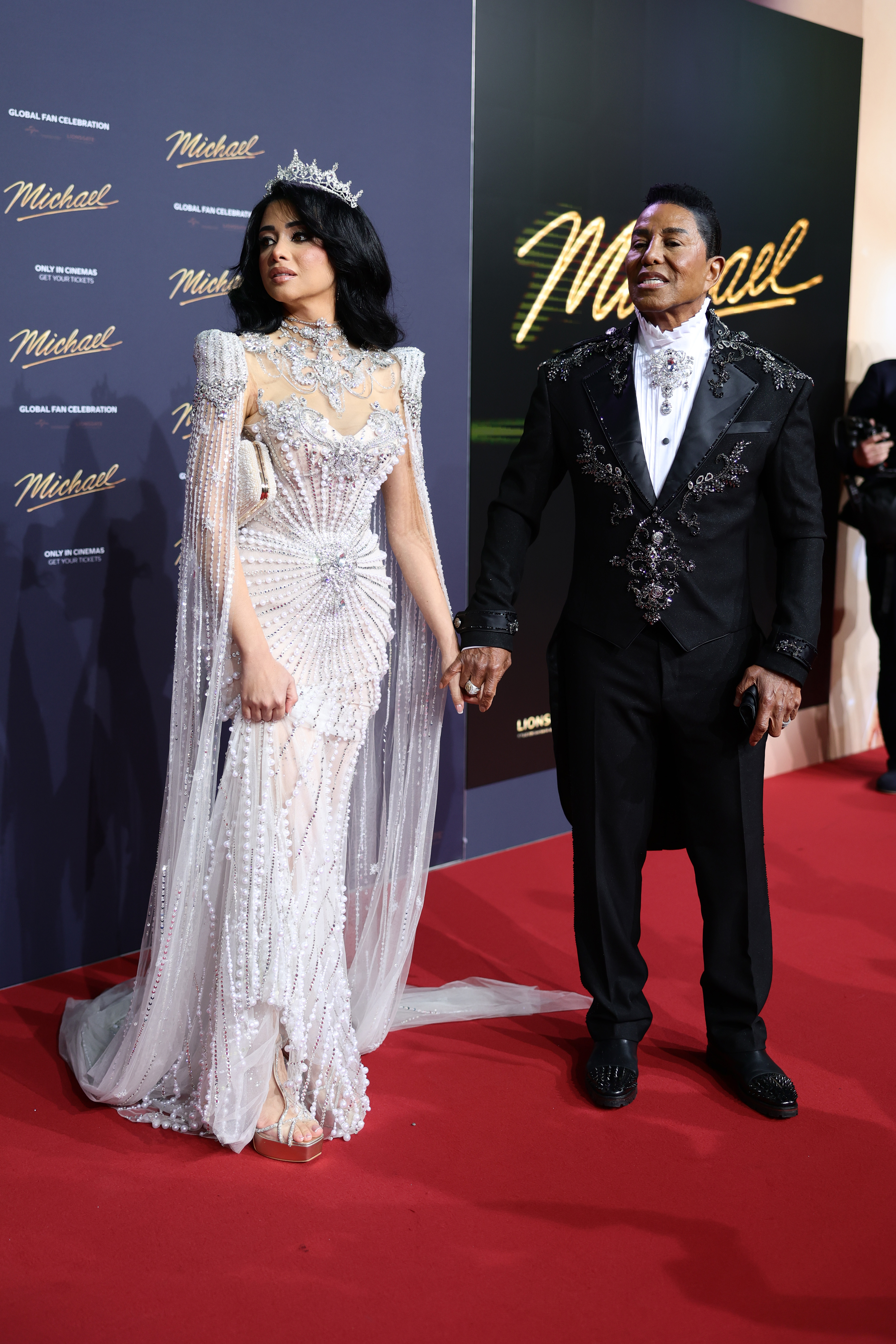 Jermaine Jackson and his wife pose together on the red carpet at the Berlin premiere, presenting a striking, coordinated look. Her ornate, crystal-embellished gown and tiara contrast elegantly with his detailed black suit, creating a dramatic, high-glamour pairing.