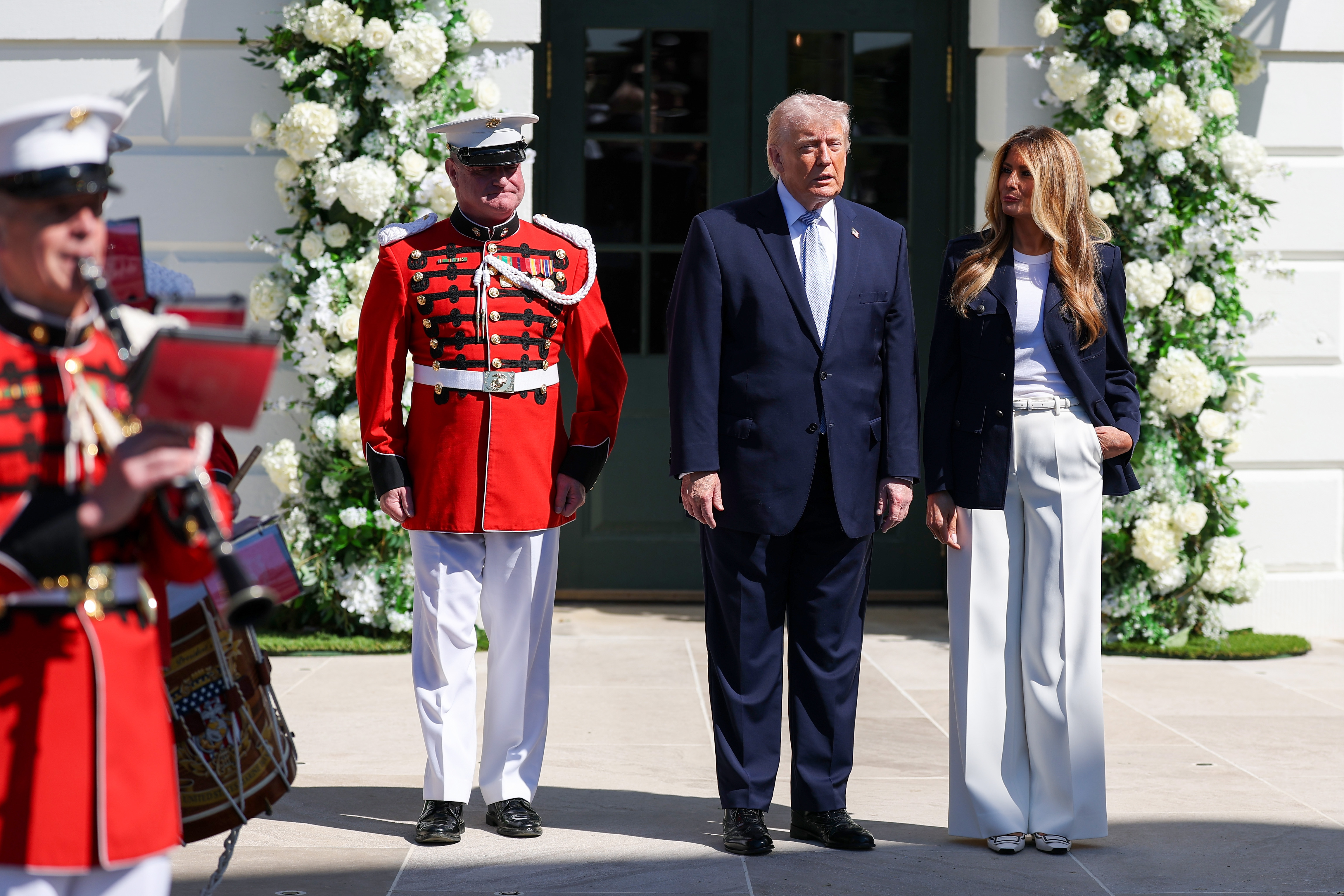 Donald and Melania Trump stand beside a uniformed band member near the flower-framed White House entrance as they greet guests, continuing a tradition that began in 1878.