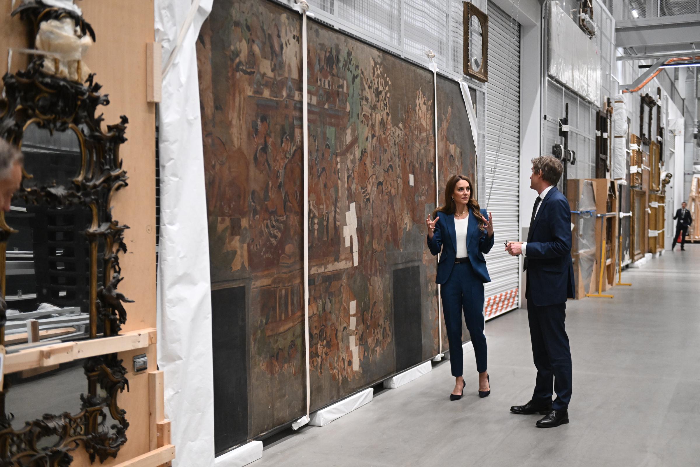 In the storage hall, Princess Catherine speaks with V&A Director Tristram Hunt in front of a large mural. Her hands raised in conversation, she appears animated and attentive, dressed in a sharp blue suit and suede heels.