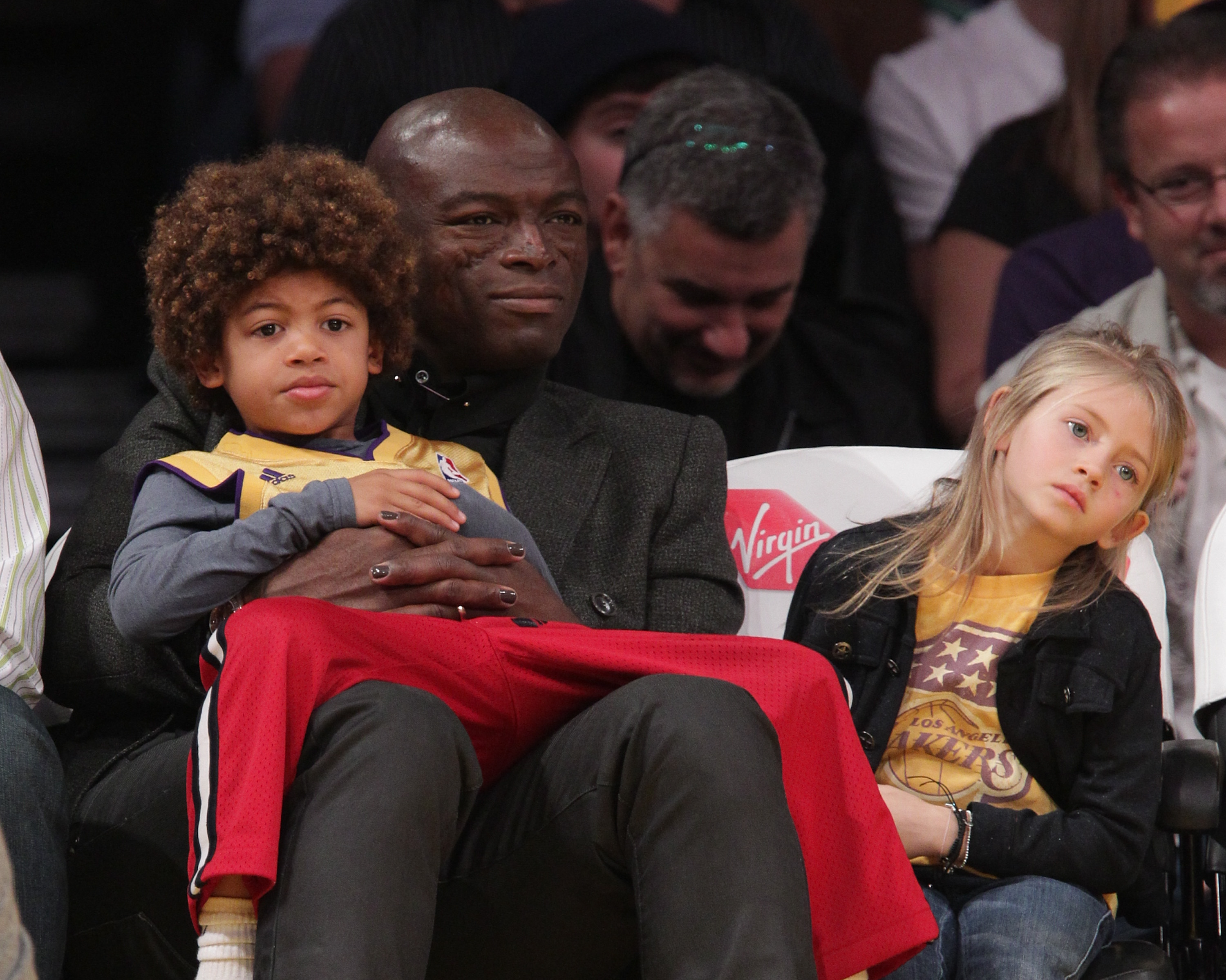 Courtside under bright arena lights in Los Angeles, Seal sits calmly with young Henry Samuel nestled in his arms, while Leni leans beside them.