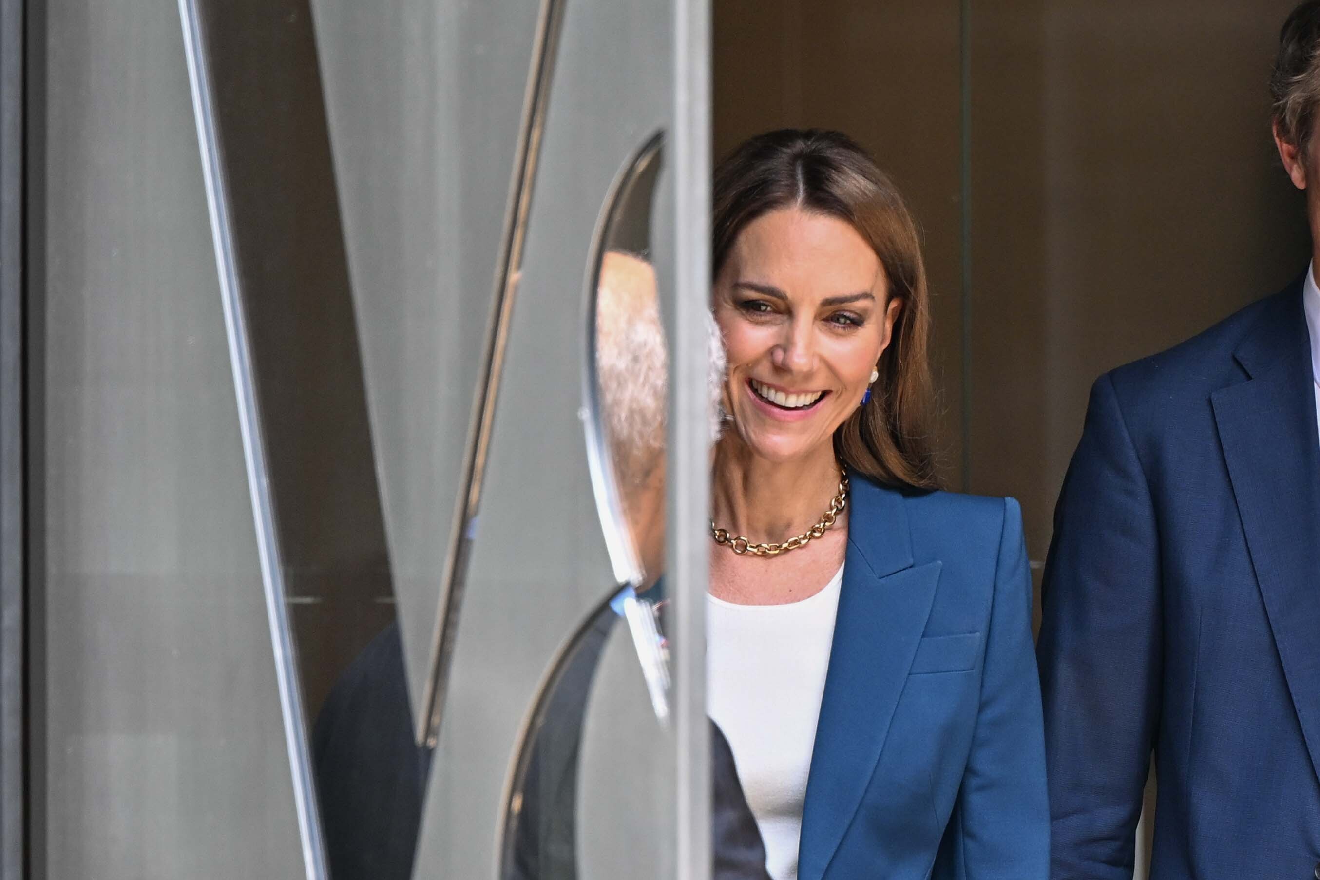 Princess Catherine shares a bright smile near the entrance of the V&A East Storehouse. She wears a tailored blue suit and white top, with gold and sapphire jewelry adding a refined touch to her modern, understated look.