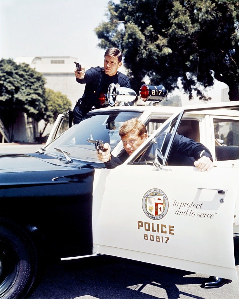 Martin Milner, US actor, and Kent McCord, US actor, both in costume as LAPD police officers, pointing handguns as they shield beside their police car in an image issued as publicity for the US television series, 'Adam-12', USA, circa 1972 | Source: Getty Images