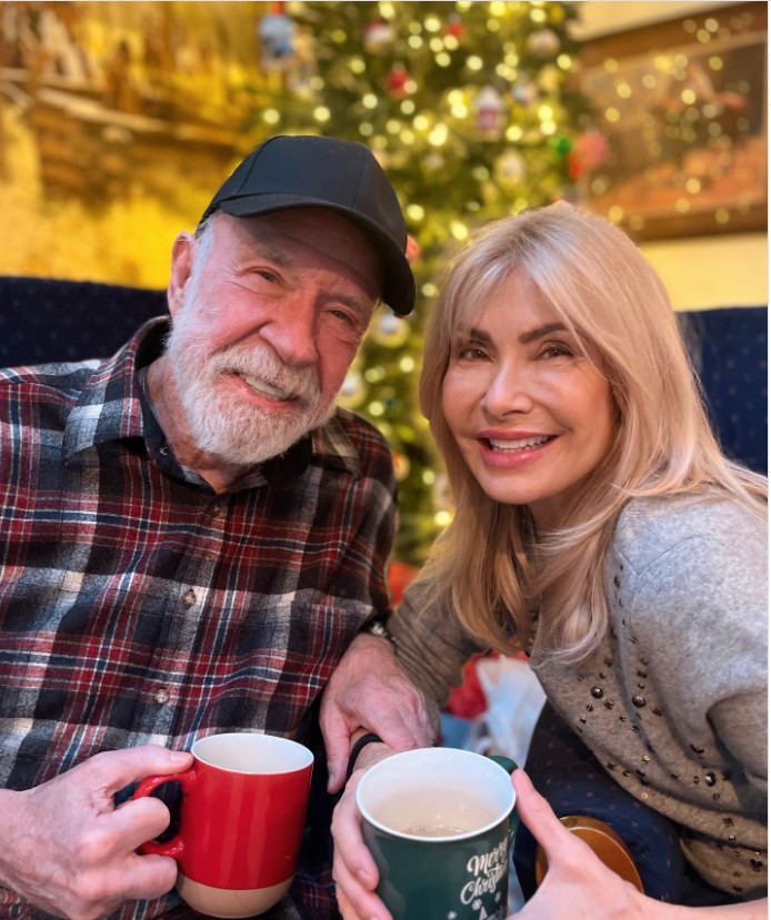 Chuck Norris and his wife Gena sit close together, smiling warmly as they hold festive mugs in front of a glowing Christmas tree. | Source: Instagram/chucknorris