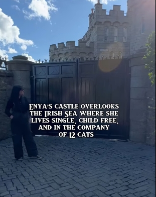 Standing before the tall, dark gates of her castle, a lone figure gazes up at the stone façade, dwarfed by its fortified walls and sunlit towers. The scene underscores the scale and privacy of the residence Enya made her own, a home designed as much for retreat as for creative reflection. | Source: YouTube/@beforethecoffin