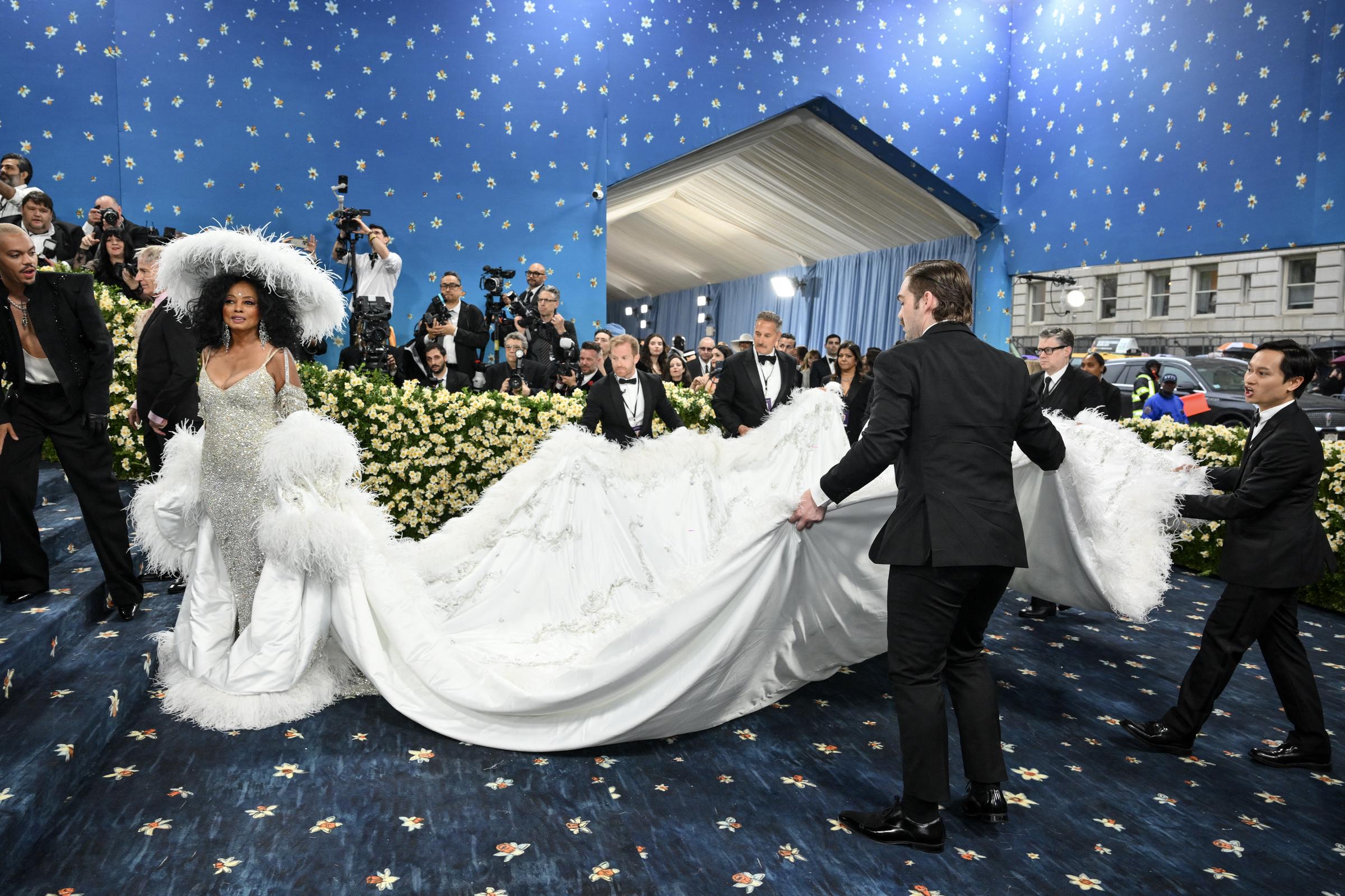 In this moment, Ross stands poised at the base of the stairs as a team of assistants carefully lift and arrange her voluminous train. The gown fans out around her, its embroidery and delicate textures catching the light.