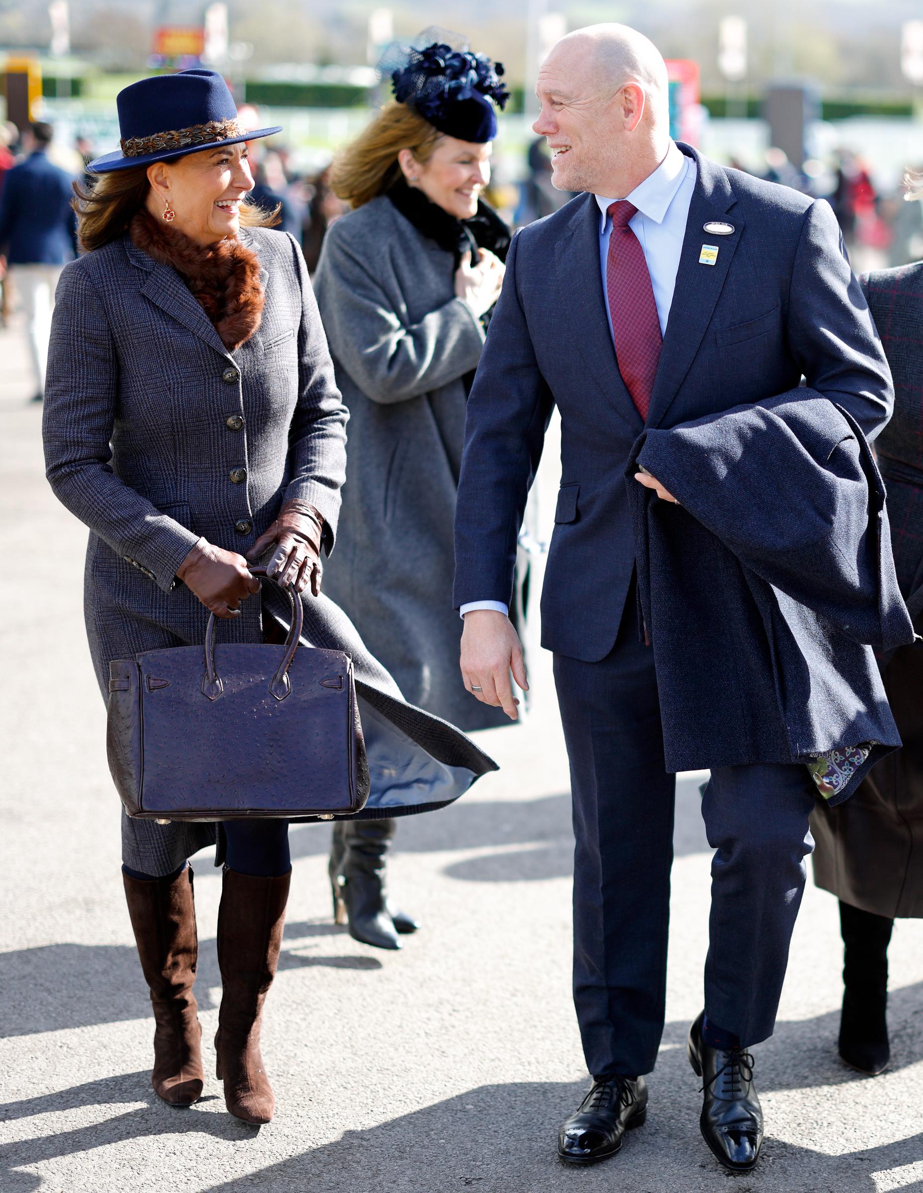 Carole Middleton shares a laugh with Mike Tindall — husband of Zara Tindall and Prince William's cousin by marriage — as the pair stroll side by side through the racecourse crowds, Carole's dark Hermès Birkin bag gripped in her gloved hands and her caramel faux fur scarf catching the breeze, the easy warmth between them suggesting the two are very much old friends by now.