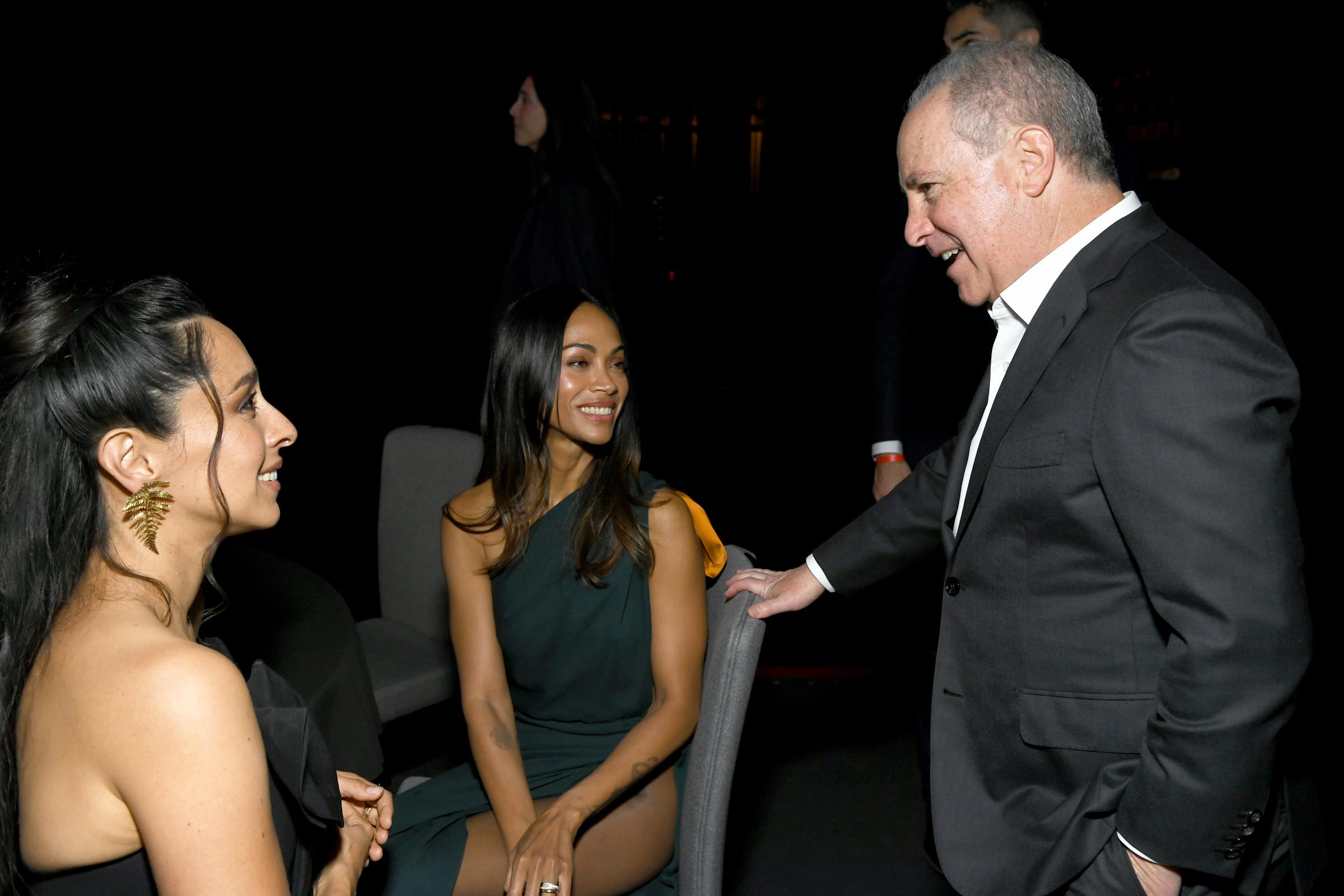 Zoe Saldaña is seated between Oona Chaplin and Alan Bergman, sharing a smile in conversation. The candid moment captures a warm exchange during the premiere event.