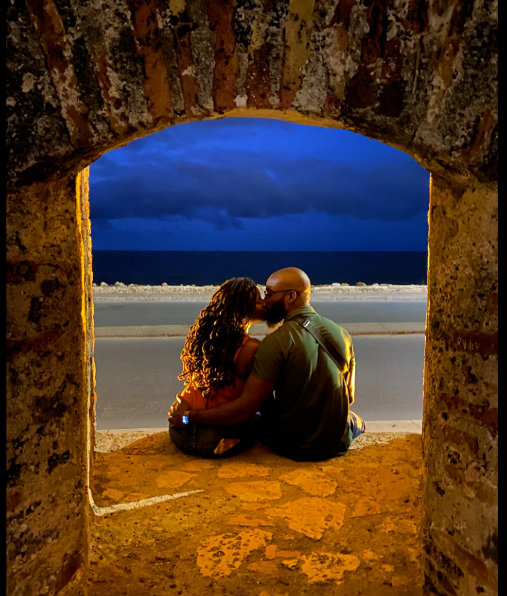 Framed by a rustic stone archway, Nancy Metayer Bowen and her husband Stephen Bowen share a quiet kiss by the seaside, the glowing evening light and deep blue horizon creating a strikingly intimate, cinematic moment. | Source: Facebook/Nancy Metayer Bowen