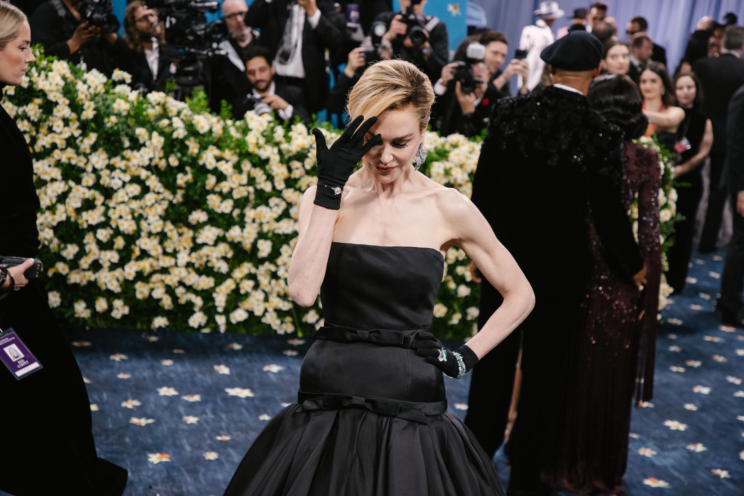 Nicole Kidman blushes softly as she strikes a playful pose for the camera, one hand gently brushing through her cropped hair while the other rests on her waist. The moment captures a rare mix of elegance and ease under the Met Gala spotlight.