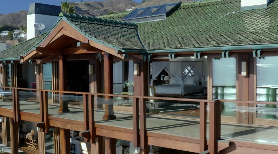 An exterior shot showcases the second-story primary suite balcony, complete with glass-paneled railings, warm timber beams, and a green clay-tiled roof. Large sliding glass doors open directly into the spacious bedroom, offering an unobstructed view of the four-poster canopy bed from the outside deck. | Source: YouTube/Architectural Digest