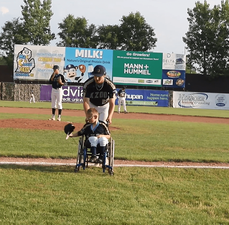 A baseball player for the Kalamazoo Growlers pushing Brenden Lowery in his wheelchair off a football field. │Source: Facebook/kzoogrowlers