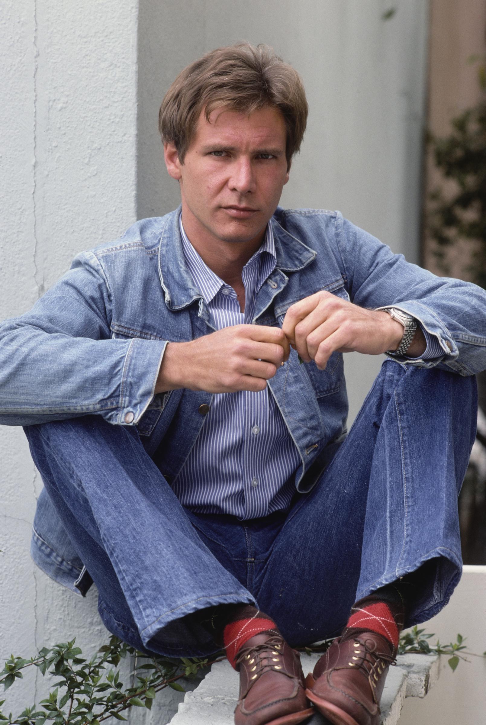 Rocking the ultimate late-1970s style, a rugged Harrison Ford strikes a casual pose in a classic denim-on-denim outfit paired with striped shirting and eye-catching red argyle socks. With his intense, direct gaze, the rising star was already proving he had the effortless cool factor required to anchor Hollywood's biggest franchises. | Source: Getty Images