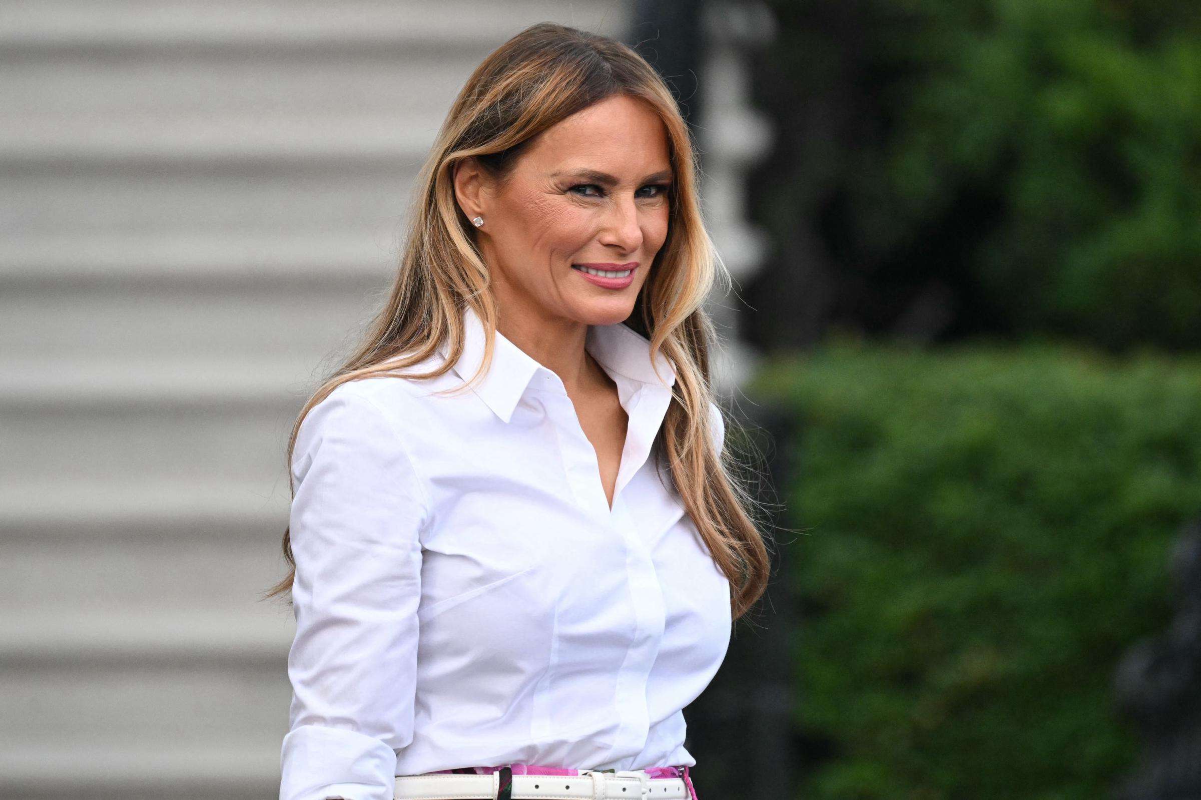 First Lady Melania Trump walks across the South Lawn of the White House during the Congressional Picnic on June 12, 2025. Dressed in a crisp white blouse and light makeup, she smiles confidently while stepping past greenery and stone steps.