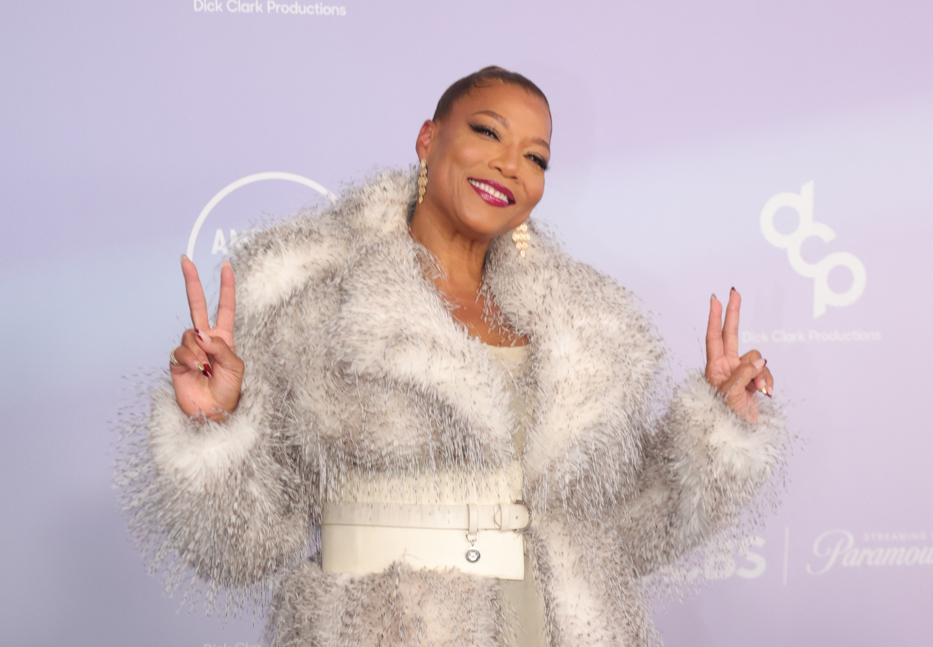 Queen Latifah flashes double peace signs and tilts toward photographers during a playful red carpet pose at the 2026 American Music Awards.