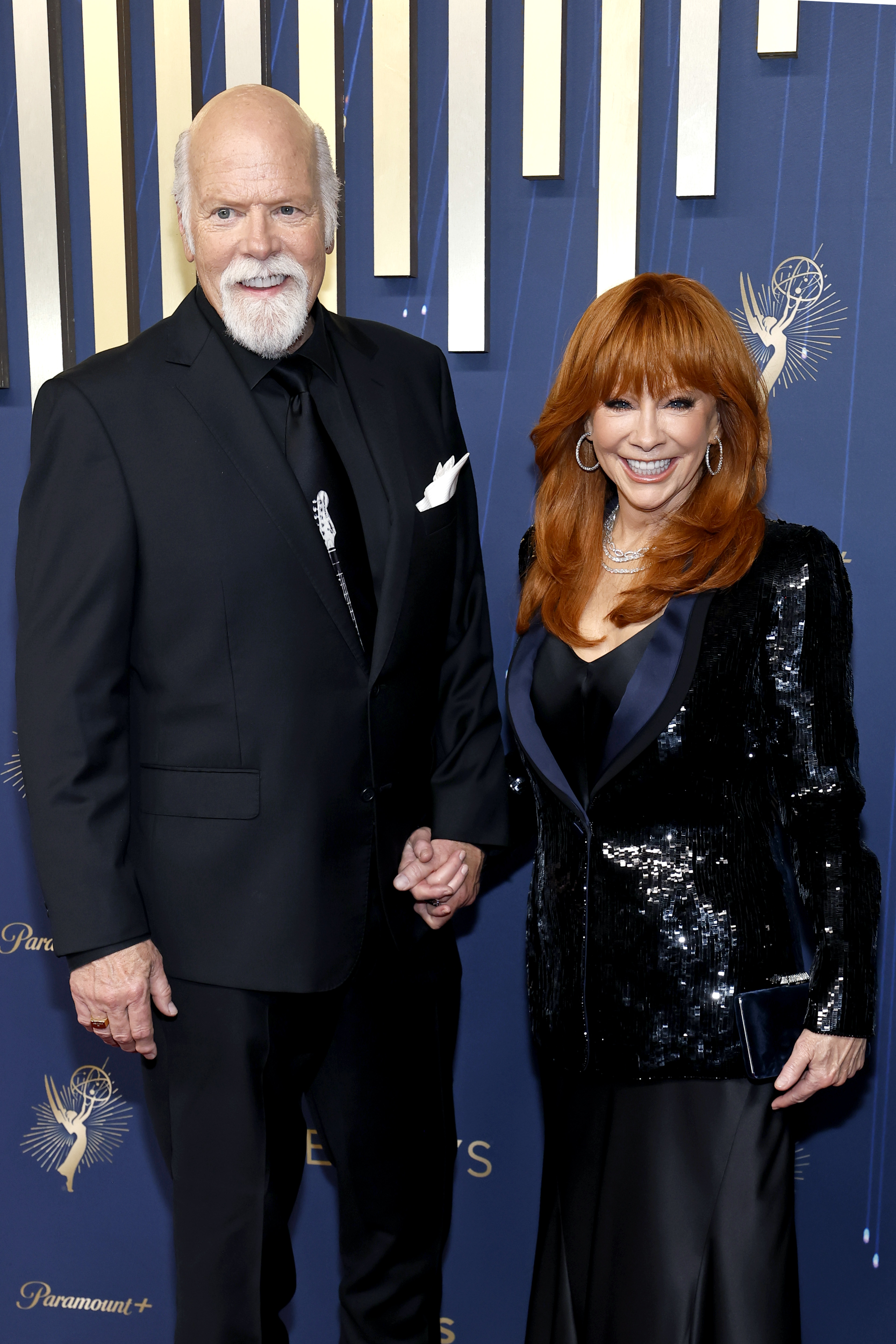 A the same event, they pose standing side by side, holding hands and smiling directly at the camera. McEntire wears a black sequined blazer over a gown, accessorized with earrings and layered necklaces. Linn wears a black suit with a pocket square.