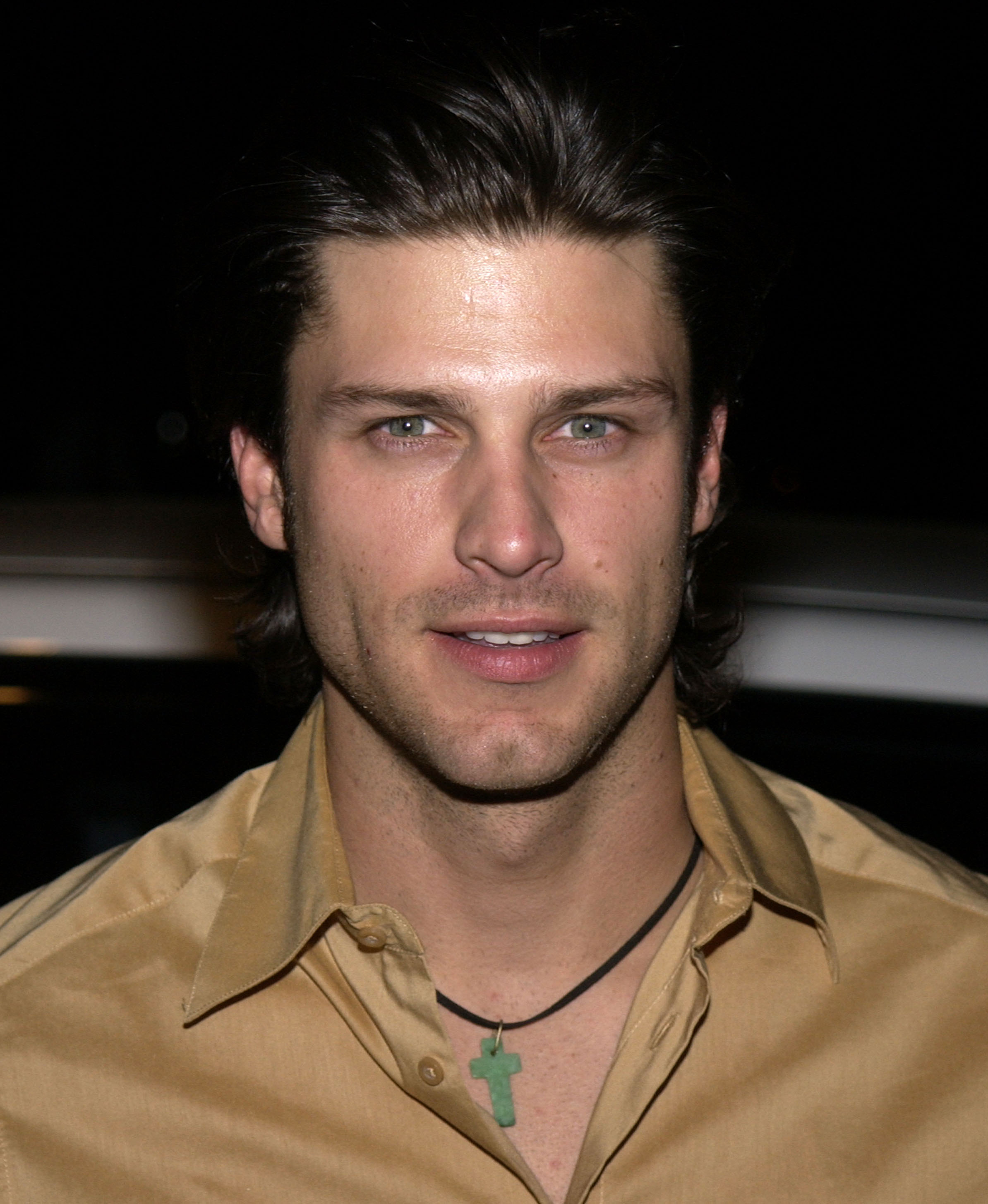 Greg Vaughan is photographed at a red carpet event in the early 2000s, wearing a gold button-up shirt and a green cross pendant necklace. His hair is swept back in a classic style, and his direct gaze, combined with the soft flash lighting, gives the image a polished yet casual feel.