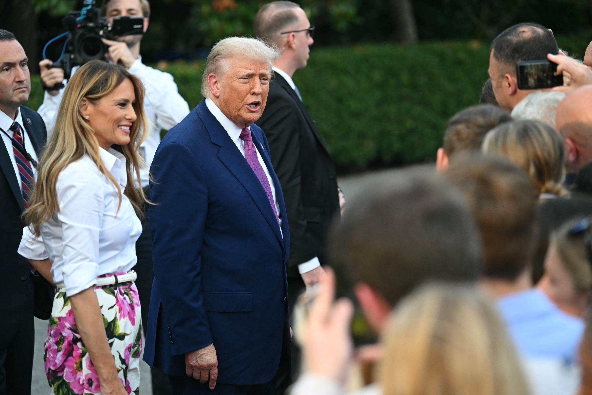 Donald and Melania Trump arrive at the Congressional Picnic, warmly engaging with attendees on the South Lawn. Melania wears floral pants and a white blouse, while Donald, in a navy suit, speaks as they are surrounded by a crowd and cameras.