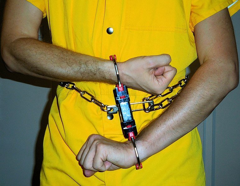 Preso en traje institucional amarillo y restricciones de alta seguridad. | Imagen: Wikimedia Commons