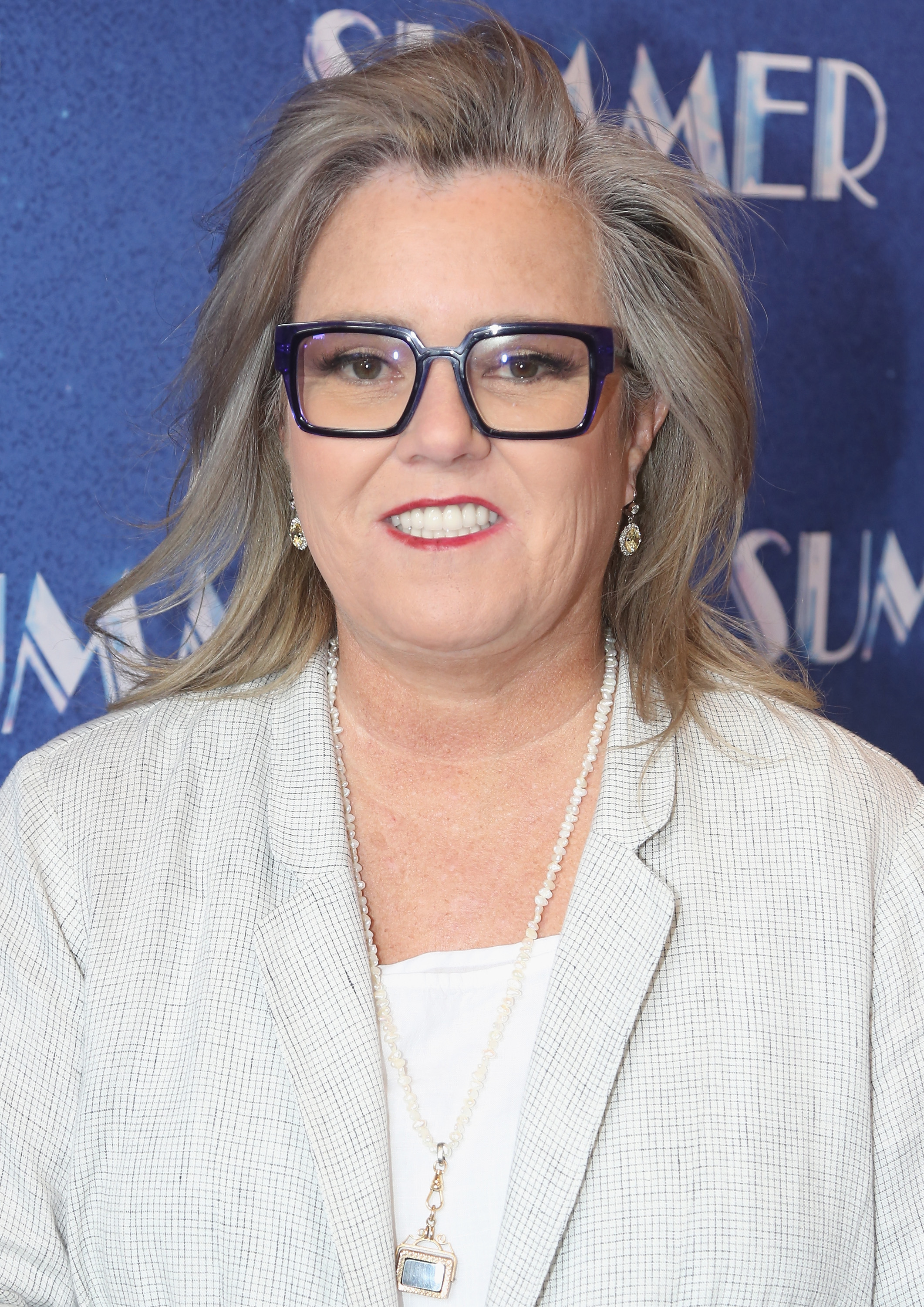 Rosie O'Donnell poses at the opening night of "Summer: The Donna Summer Musical" on Broadway, wearing a light textured blazer and layered jewelry against a blue backdrop.