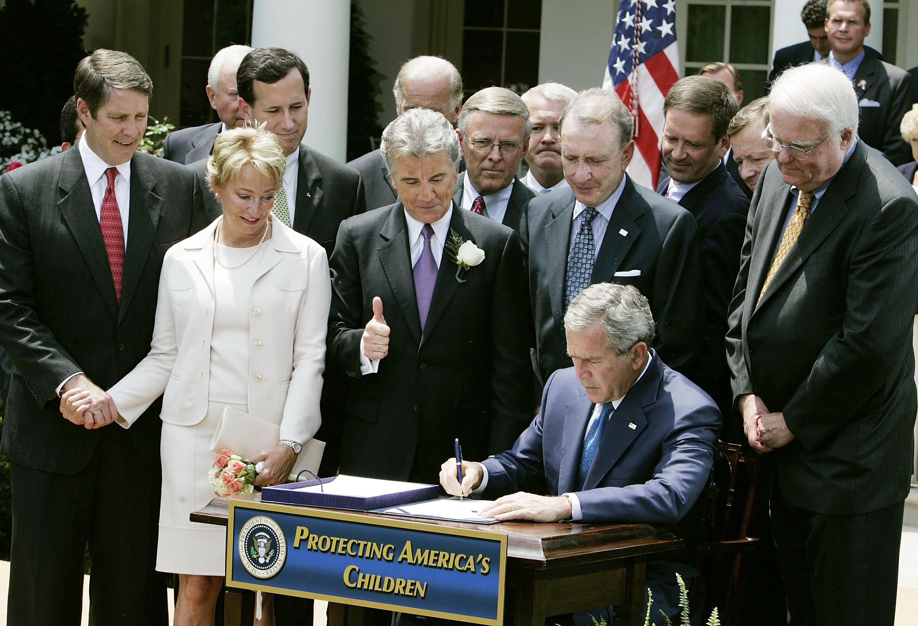 U.S. President George W. Bush signs the H.R. 4472, the Adam Walsh Child Protection and Safety Act of 2006, while John Walsh gives a thumbs up and his wife Reve Walsh (2nd-L) holds hands with Senate Majority Leader Bill Frist (L) and members of Congress stand nearby in the Rose Garden, at the White House, July 27, 2006, in Washington, DC. | Source: Getty Images