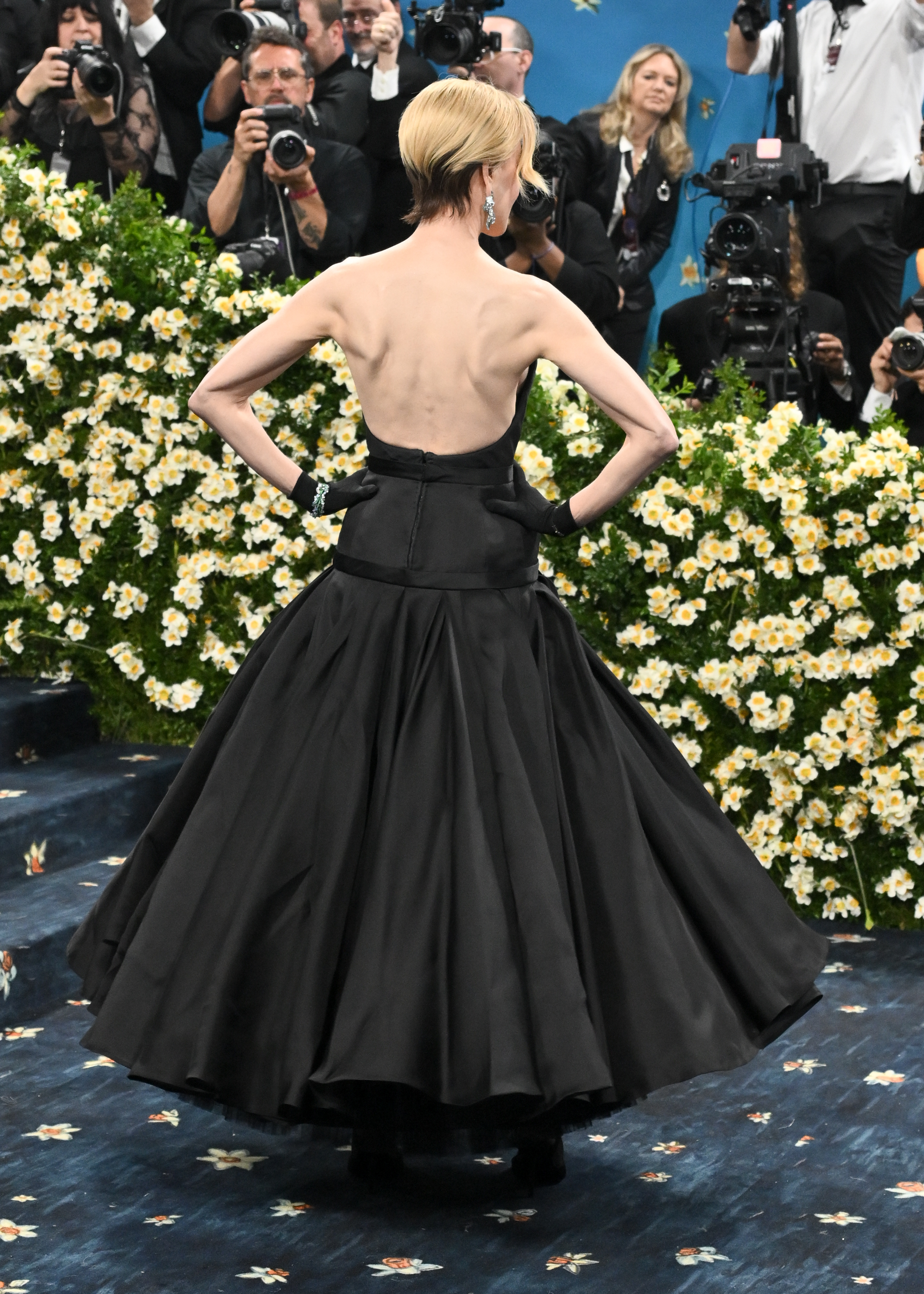 A full back shot captures Nicole Kidman as she stands tall on the Met Gala red carpet, showcasing the architectural precision of her Balenciaga gown. The minimalist design hugs her frame with elegance, while her sleek bob—platinum with dark, visible roots—adds a striking contrast.