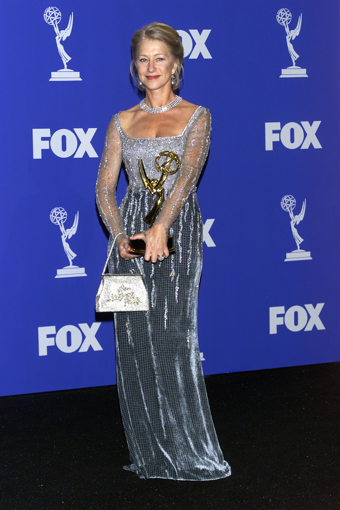 Helen Mirren shines at the Emmy Awards, holding her statuette in a floor-length silver gown adorned with shimmering beadwork, pairing the elegant look with a sparkling choker, dangling earrings, and a glittering handbag as she poses against the FOX-branded step-and-repeat backdrop.