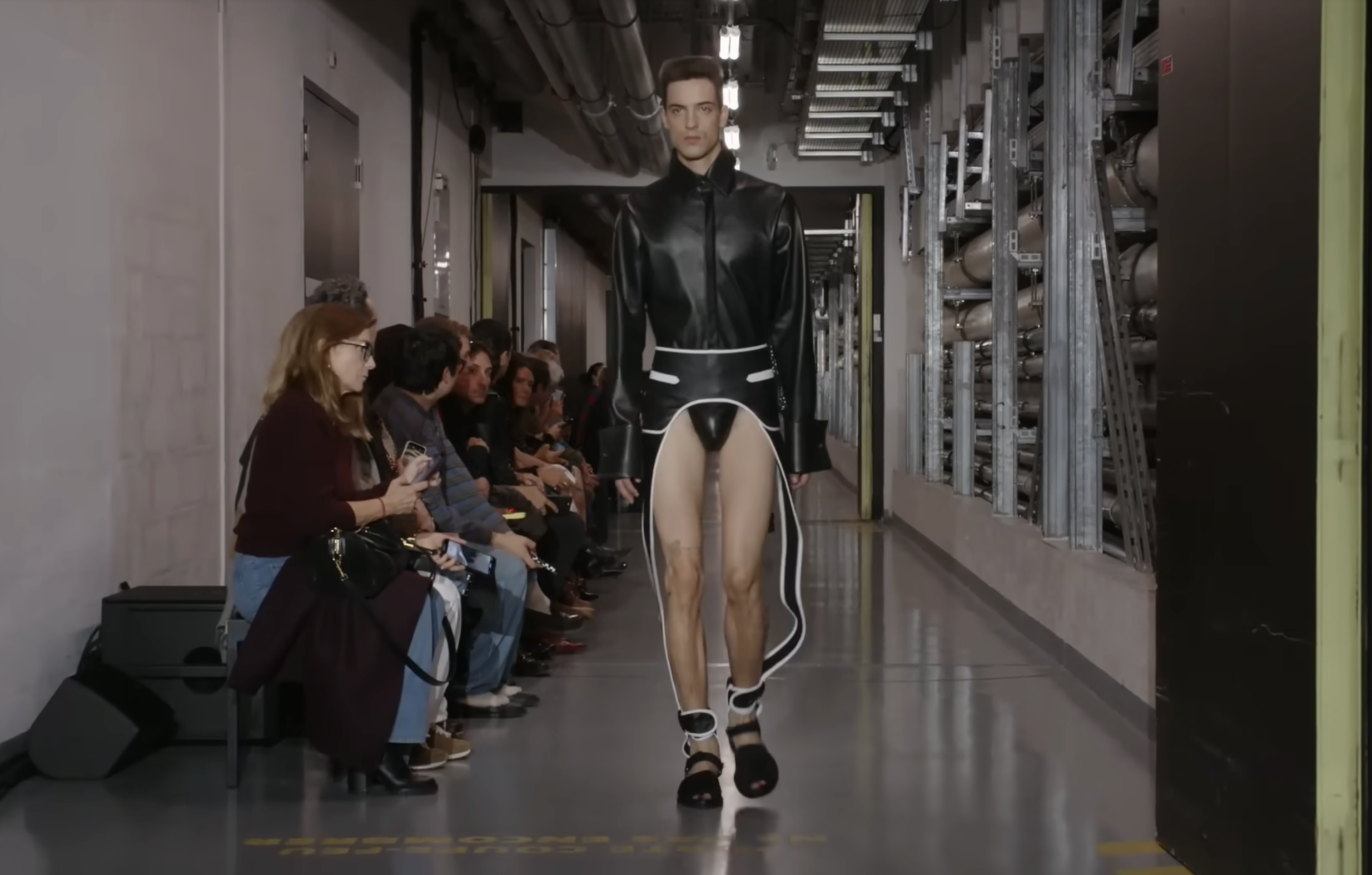 A model walks by in a striking black, groin-baring outfit. The design resembles a coat but features side detailing that extends down the legs, along with a fitted bodysuit section at the center. From a post dated October 5, 2025. Source: Youtube/@jeanpaulgaultier
