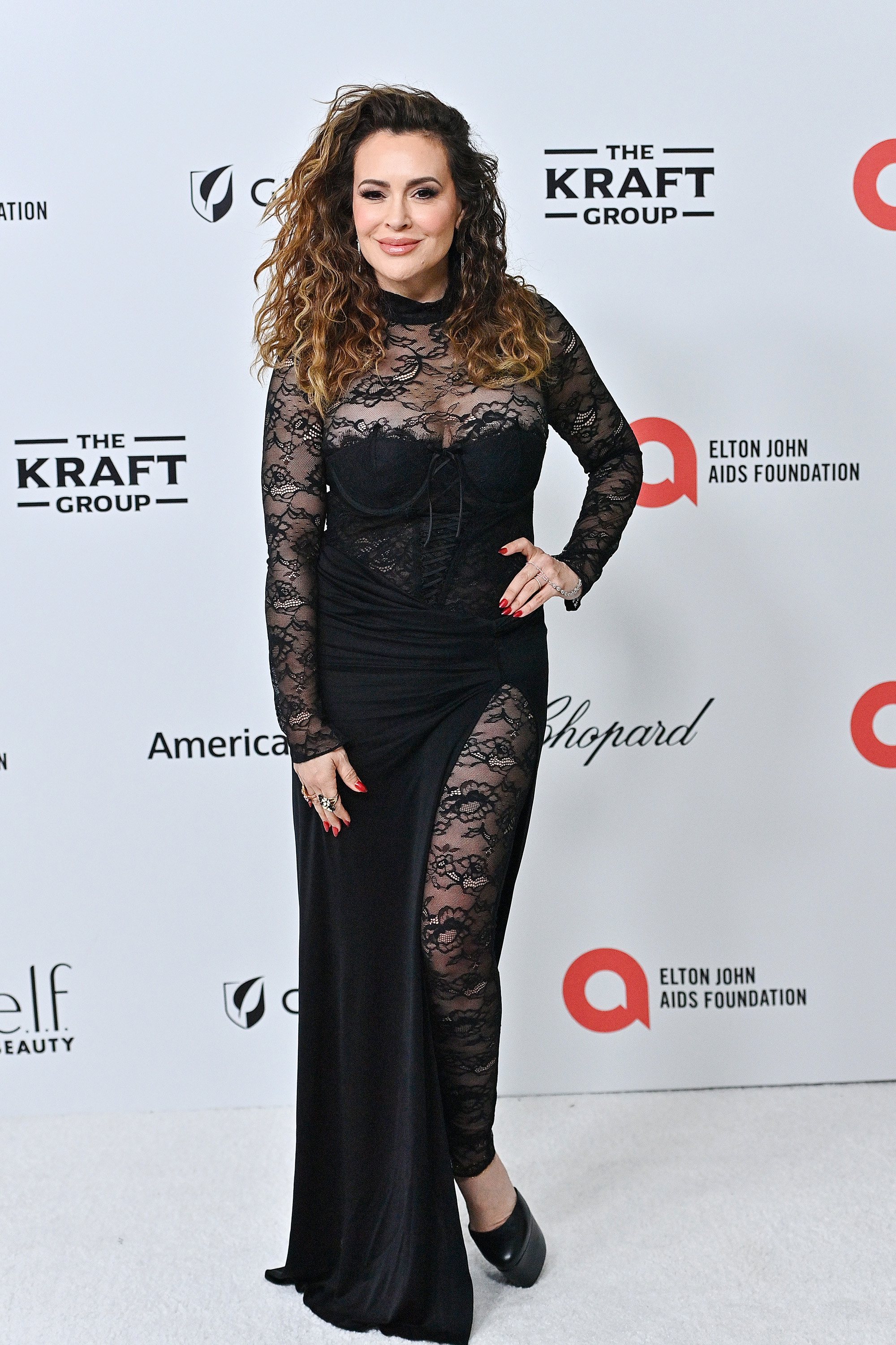 Alyssa Milano attends the Elton John AIDS Foundation event in 2024, wearing a striking black gown with sheer lace panels. Her voluminous curls, bold eye makeup, and red nails add to the dramatic elegance of the look. Posing confidently on the white carpet, she brings a sense of glamour to the high-profile occasion.