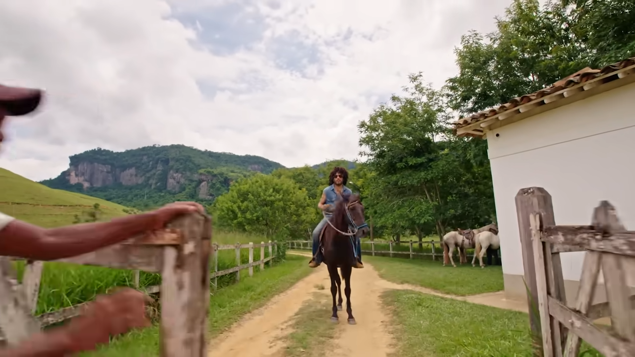 Lenny Kravitz rides a dark brown horse down a dirt path on his sprawling Brazilian estate, as a farmhand opens a rustic wooden gate. | Source: YouTube/@Archdigest