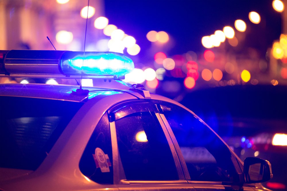 The blue light flasher atop of a police car is visible as well as city lights in the background | Photo: Shutterstock/ArtOlympic