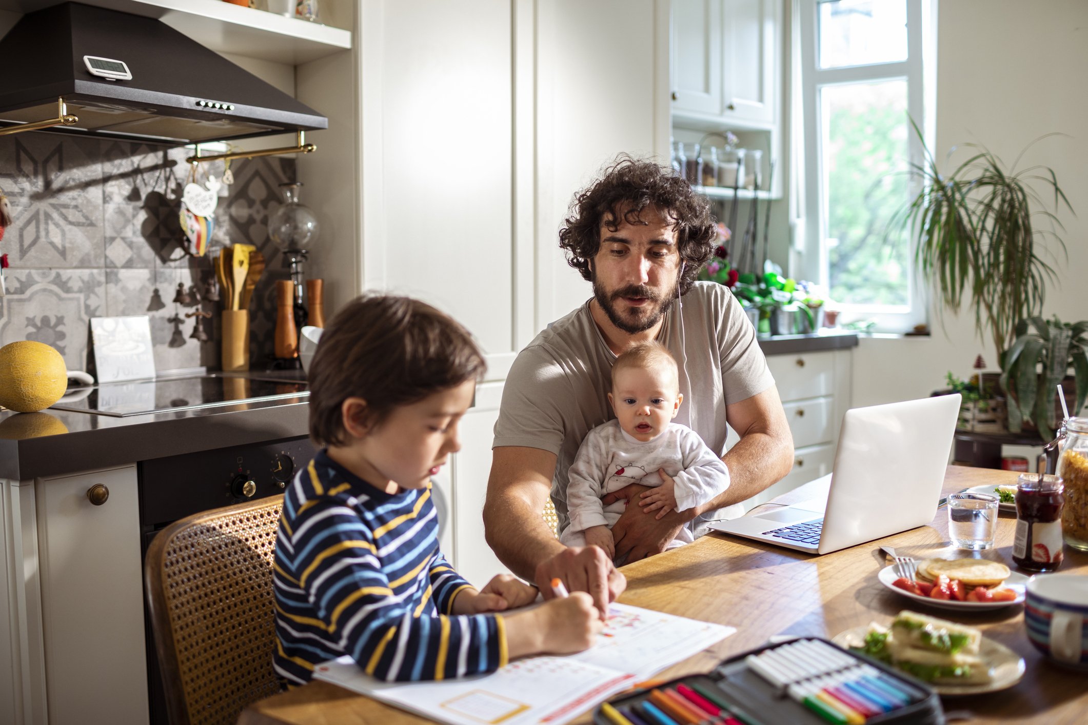 A father working from home with his son studying next to him while holding his baby | Source: Getty Images