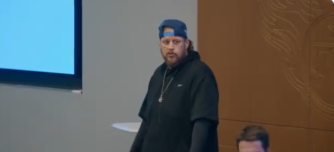 Jason "Jelly Roll" DeFord is pictured standing in what appears to be a conference or meeting room, wearing a navy blue hoodie and a blue Tennessee Titans baseball cap worn backward. His tattoos are visible on the side of his face, and he accessorizes with a silver pendant necklace. He gestures with his hands, suggesting he is speaking or making a point during a conversation or presentation | Source: X/TennesseeTitans