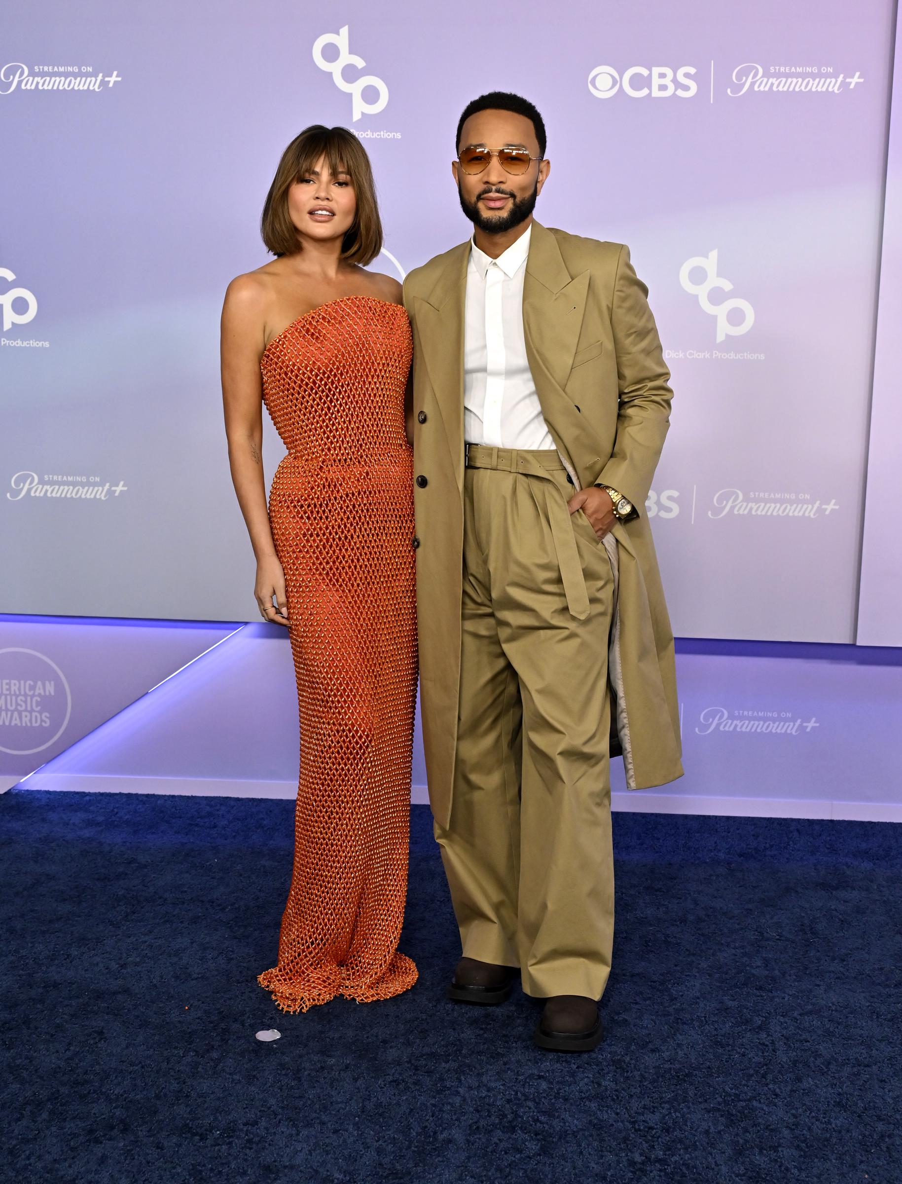 Chrissy Teigen shimmered in a figure-hugging burnt-orange gown covered in metallic beadwork that created a glowing, liquid-like effect under the lights, while John Legend complemented her in an oversized camel-toned ensemble with wide-leg trousers and a structured trench coat.