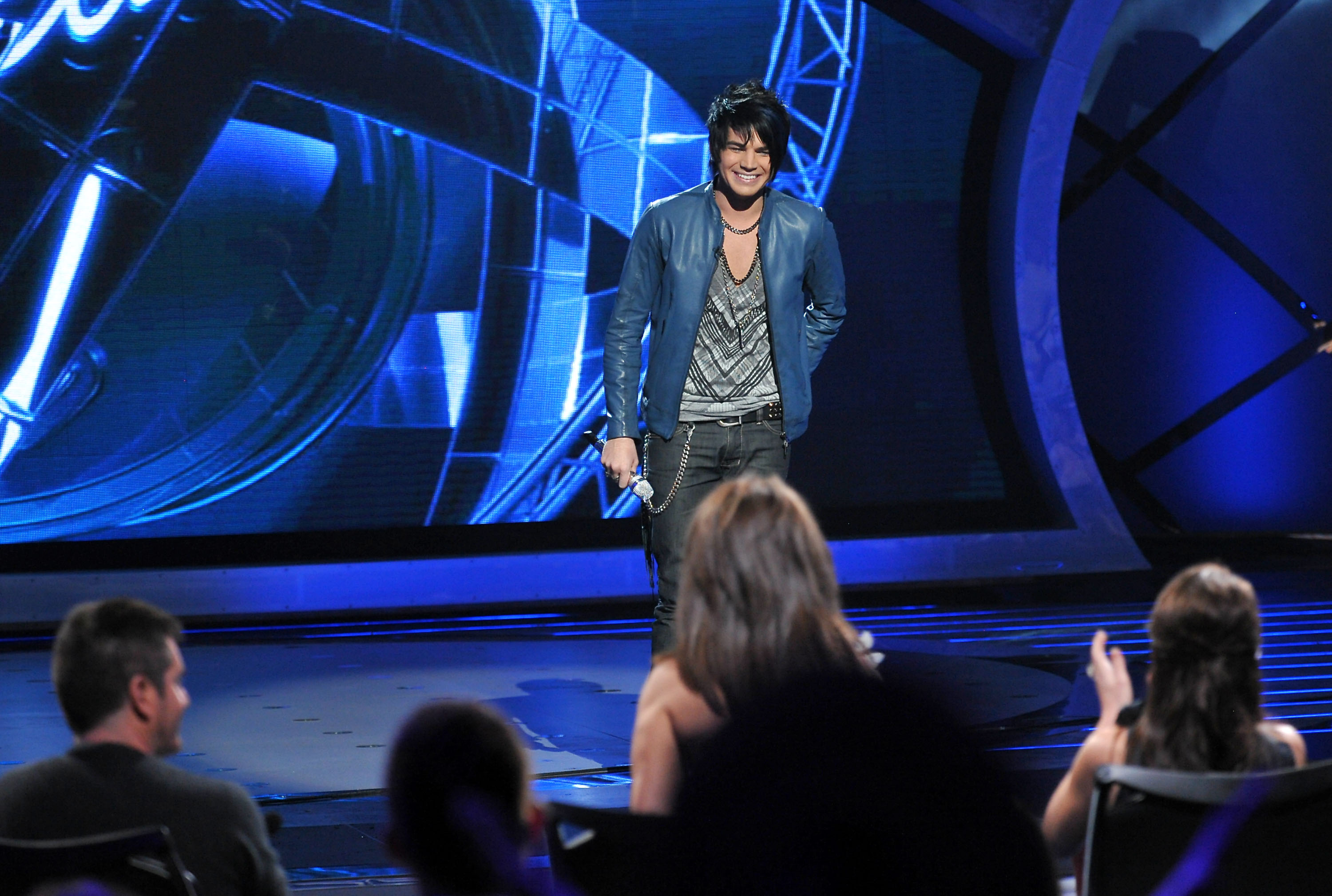 On March 10, 2009, Adam Lambert takes the "American Idol" stage in a blue leather jacket and graphic tee. With a microphone in hand and confidence in his stride, he performs live before the judges during the Top 13 round, capturing audience attention.