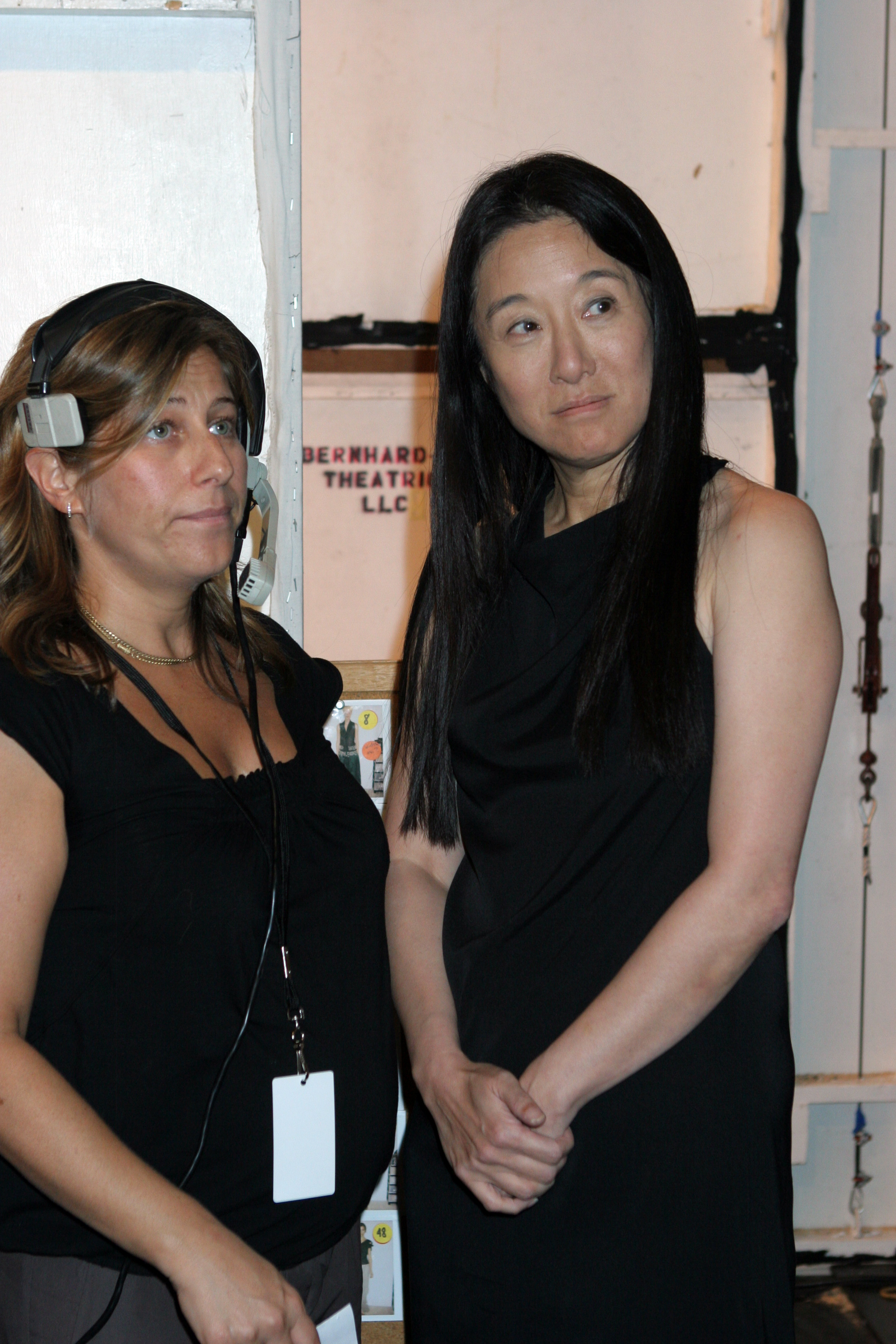 Backstage at her Spring 2006 runway show, Vera Wang stands beside a crew member, wearing a sleeveless black dress and her hair styled naturally straight. Captured in a quiet moment, she appears composed while overseeing final show details behind the scenes.