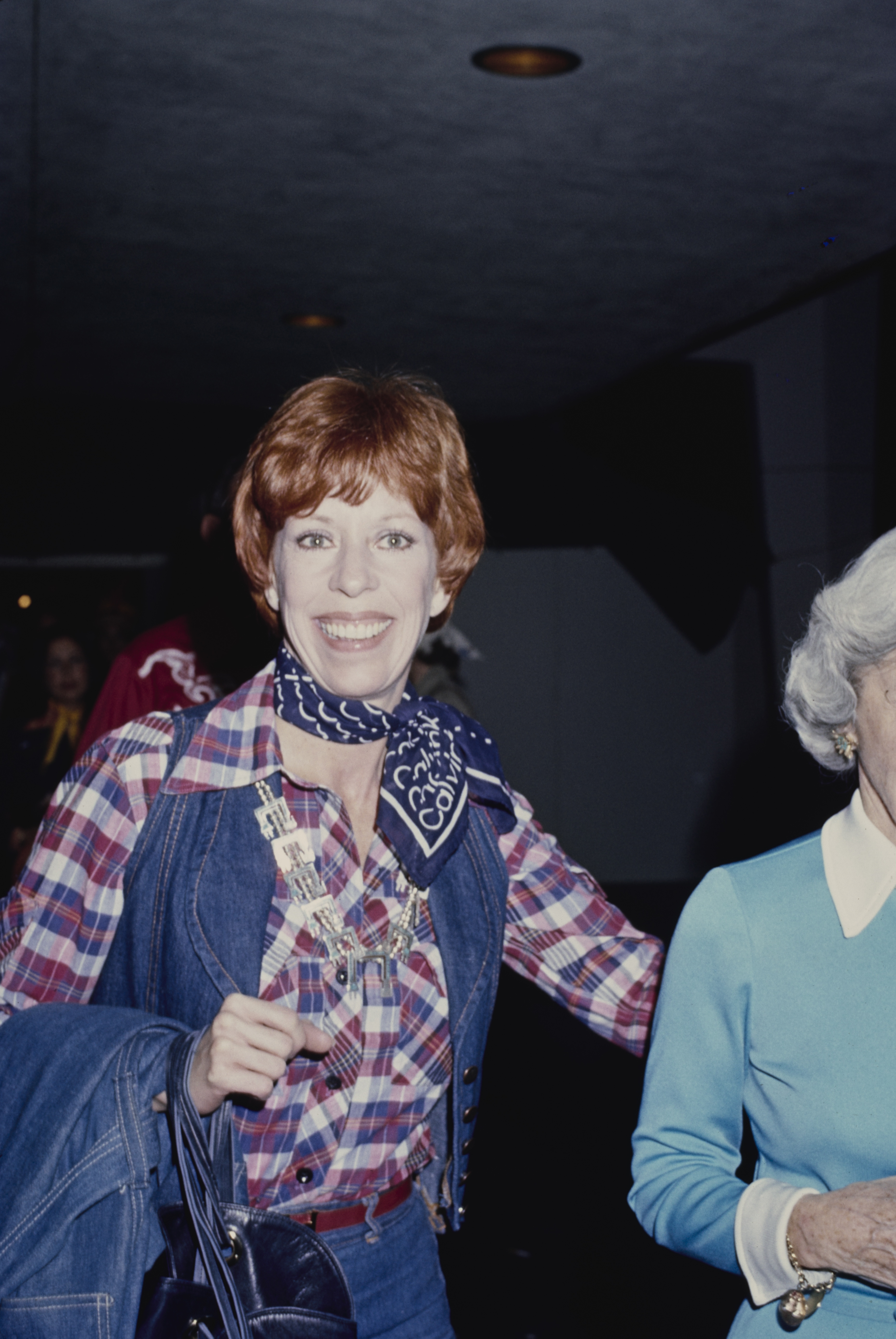Dressed in a red, white, and blue checked shirt layered under a denim vest, she adds a neckerchief tied around her neck. She carries a handbag over her arm and appears mid-step, smiling toward the camera.
