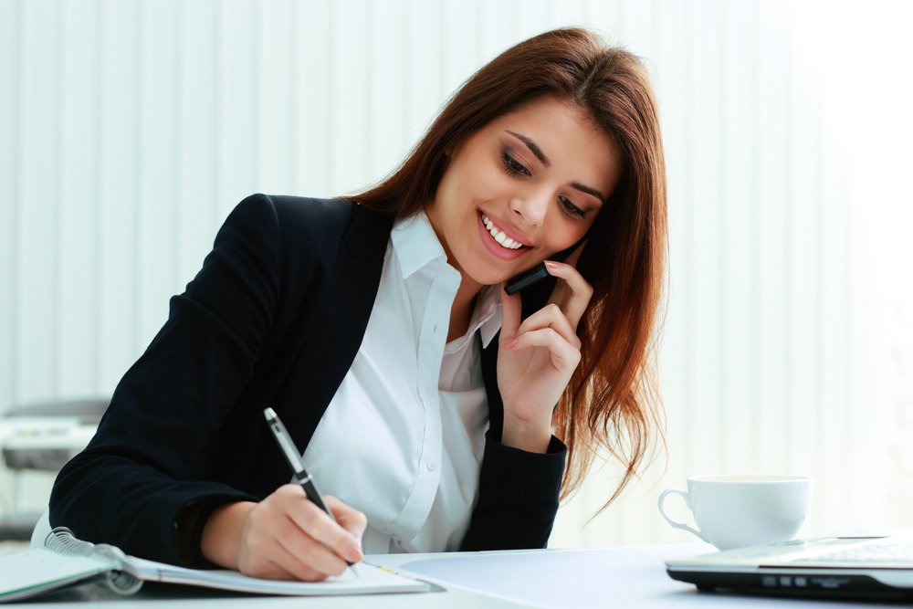 Junge, glückliche Geschäftsfrau telefoniert und schreibt im Büro Notizen. I Quelle: Shutterstock