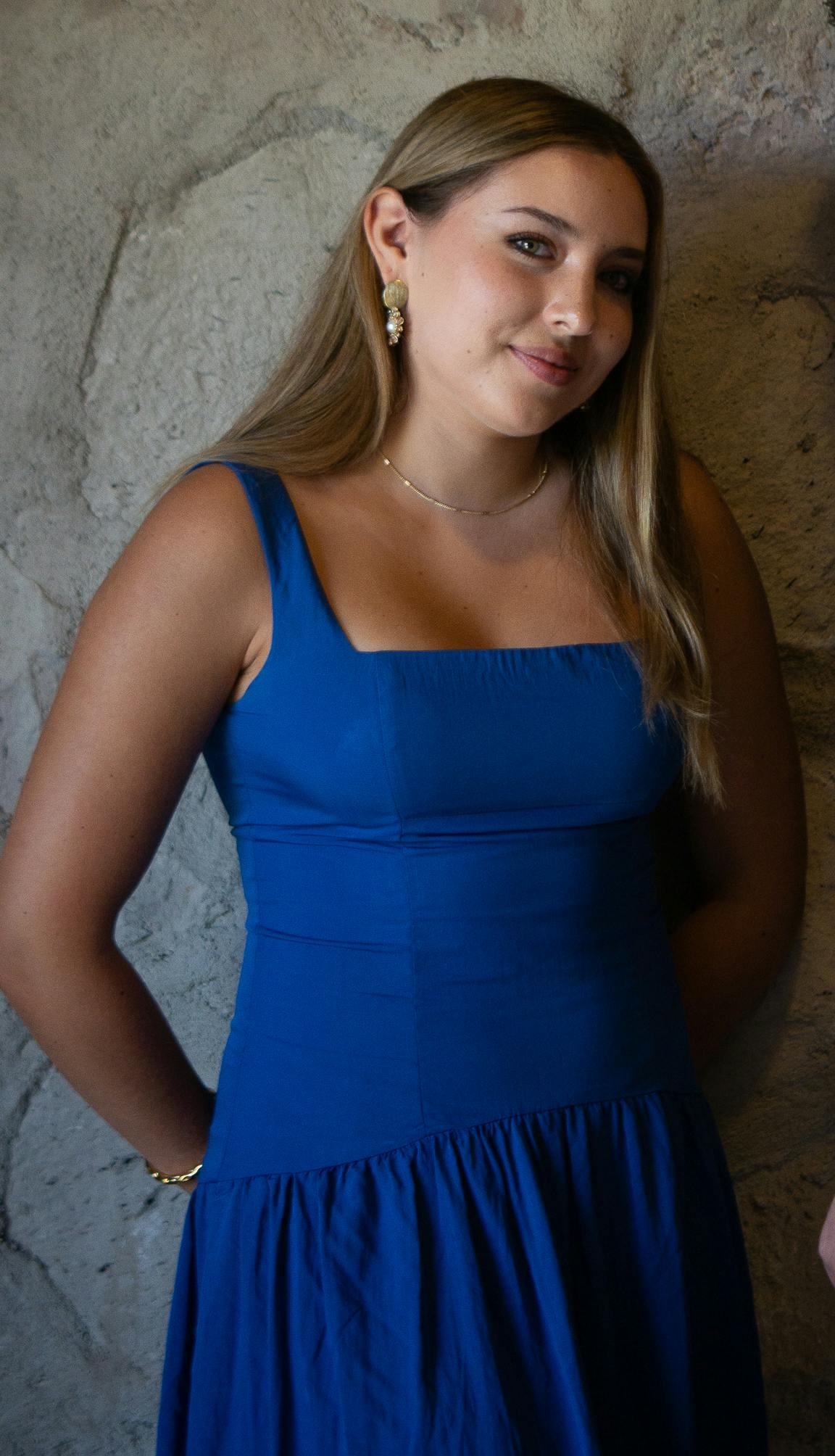Una joven sonriente con un vestido azul, collar y pendientes, de pie frente a un muro de piedra | Fuente: Pexels