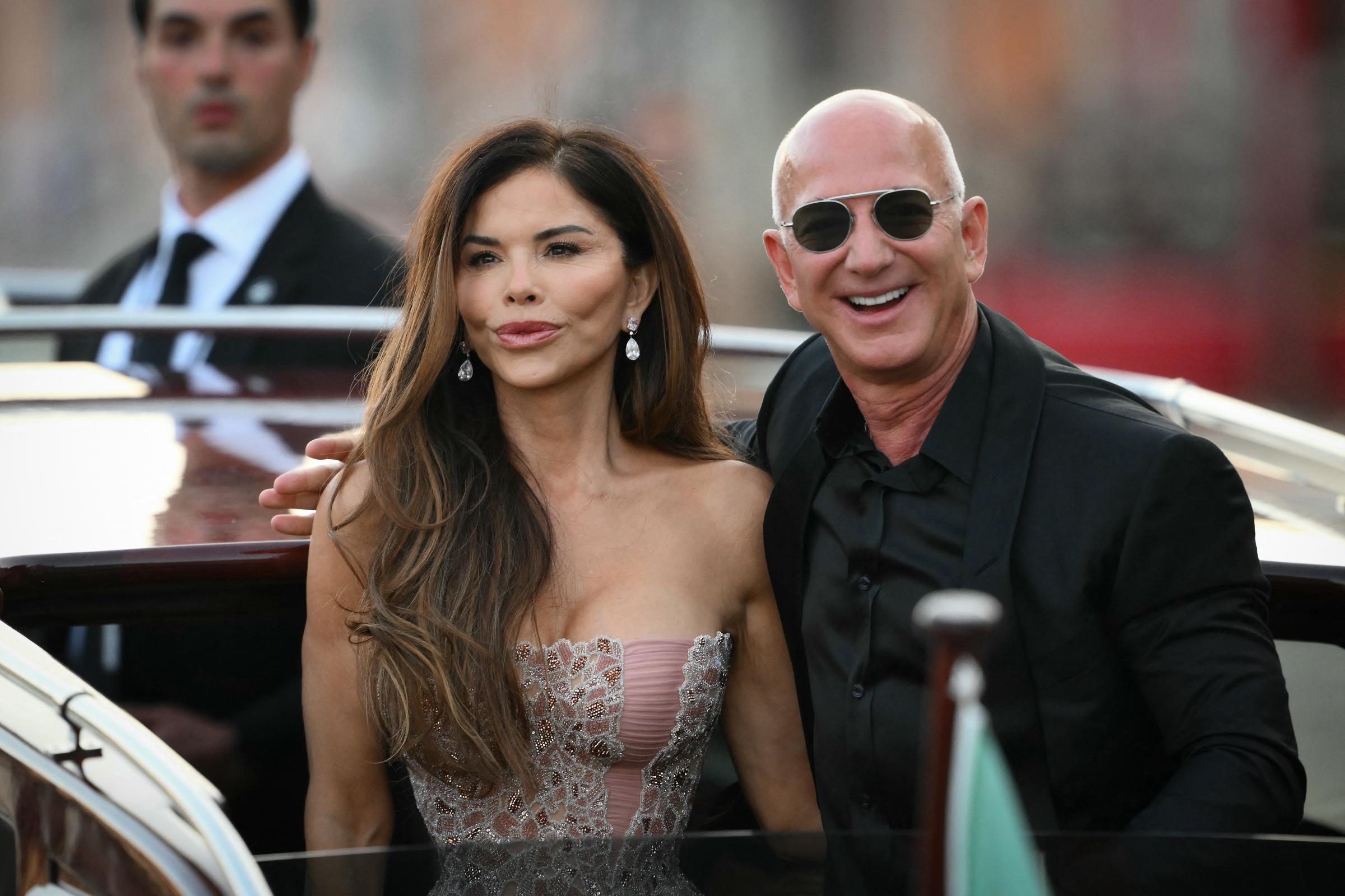 Jeff Bezos flashes a wide smile as he leans close to Lauren Sánchez, who gazes ahead with a composed expression. The couple stands on the boat’s deck, exuding confidence as a suited security guard watches from behind against a blurred cityscape.