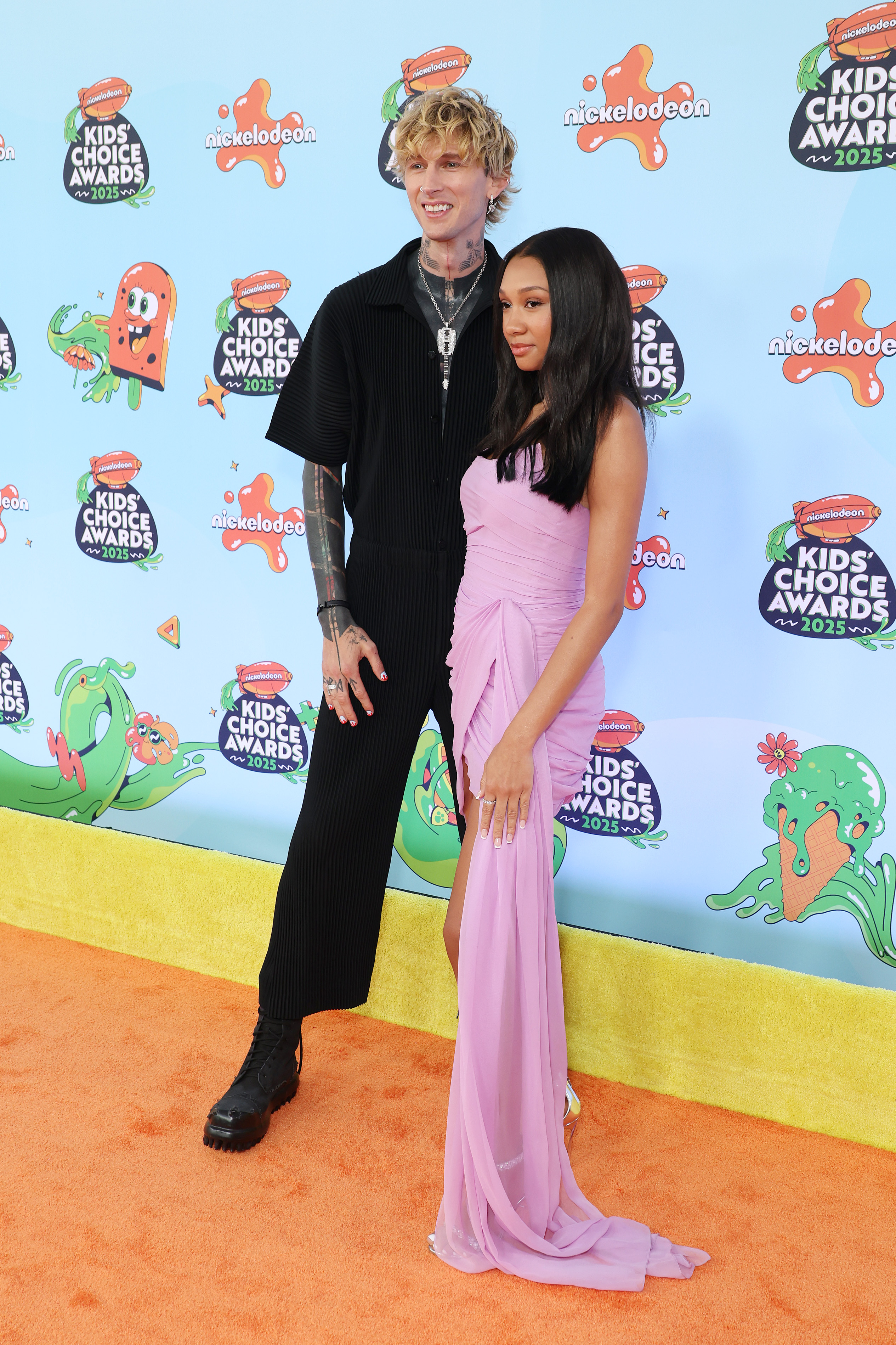Side by side, Machine Gun Kelly and Casie Baker pose with relaxed smiles and refined style. Casie's confident look and MGK’s understated outfit make for a standout duo on the orange carpet.