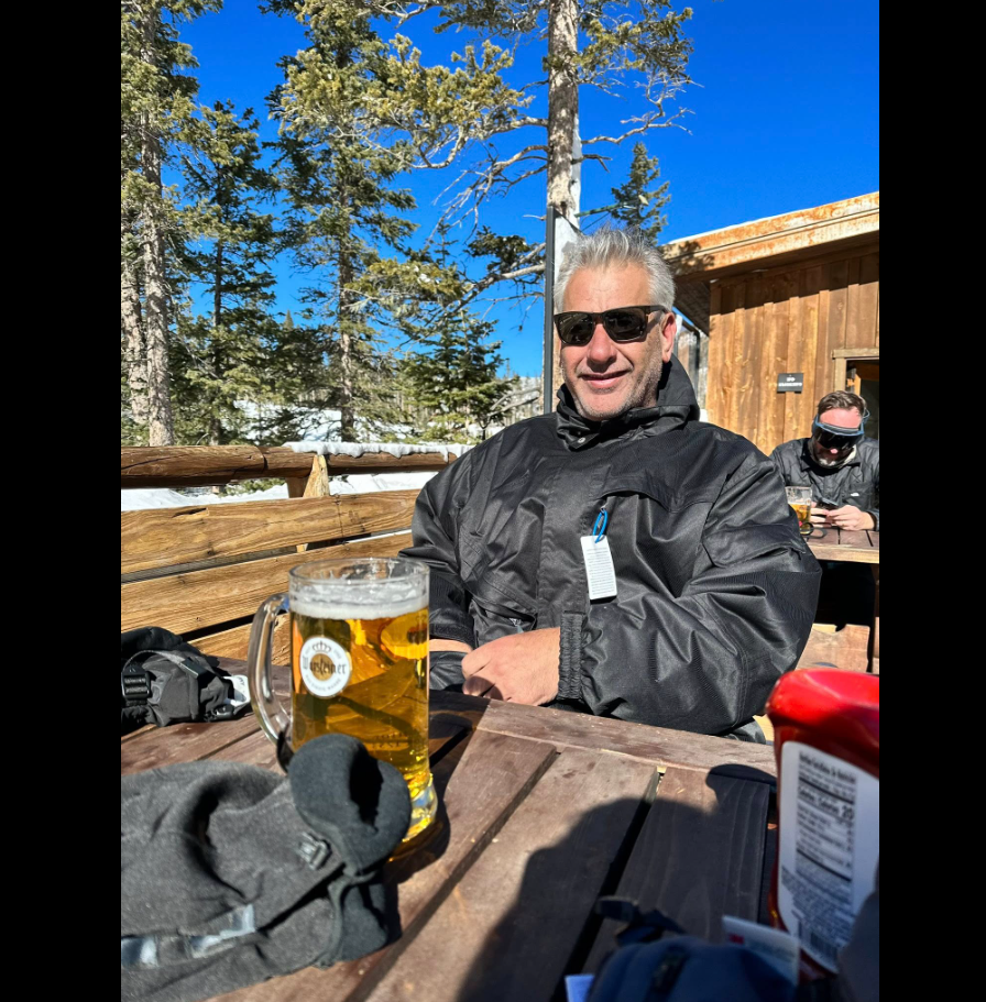 Barry Christian sits at a wooden outdoor table wearing a black jacket and sunglasses, with a glass mug of beer in front of him. | Source: Facebook/Farrah Christian