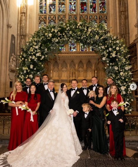 Surrounded by loved ones beneath a towering arch of white roses and greenery, Holly Ramsay and Adam Peaty posed inside Bath Abbey following their ceremony. Holly stood in the centre in a lace Elie Saab gown, holding a bouquet of white flowers, with Gordon Ramsay and Tana Ramsay close by. The image, one of nine from a post dated January 2, 2026, offered a formal yet joy-filled glimpse into the couple's wedding day. | Source: Instagram/britishvogue